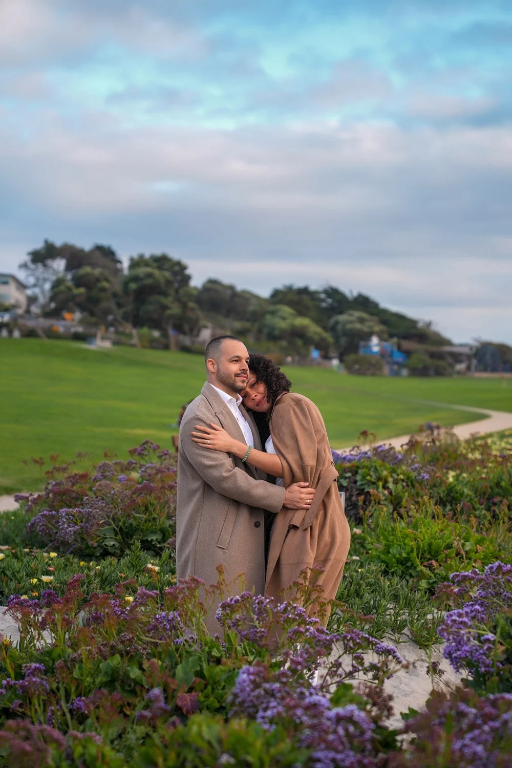 An outdoor photograph of a couple embracing amidst purple and green flowering plants on a grassy hill. The man wears a light brown overcoat, and the woman, also in a light brown coat, is leaning her head on his chest. A paved pathway winds through th