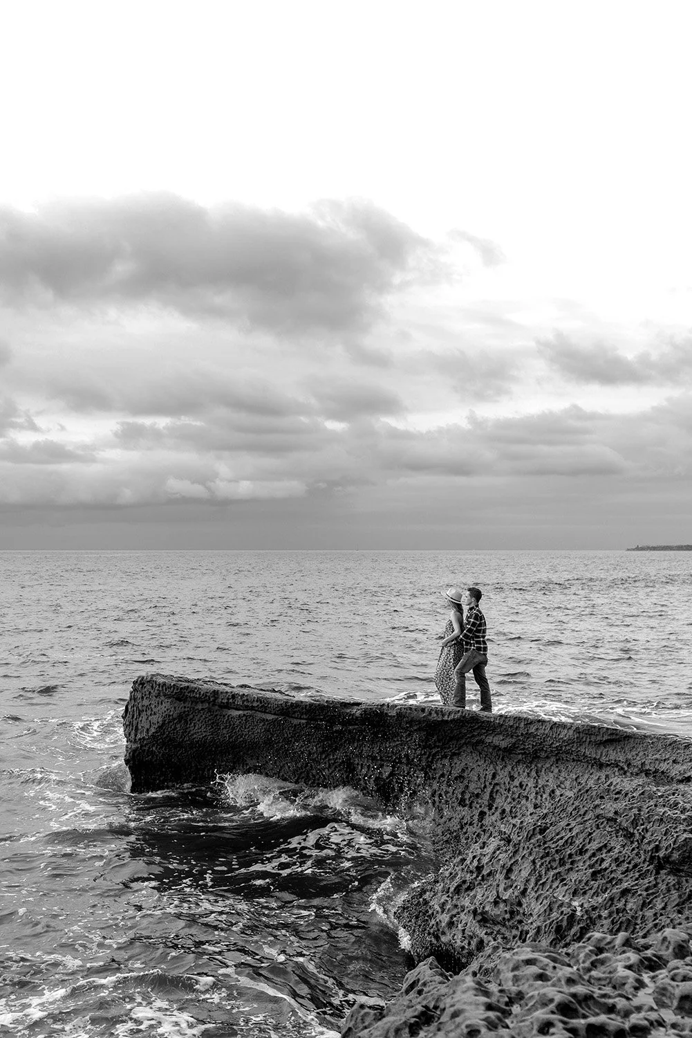 A black and white photo of a couple standing on a rock by the ocean, facing each other, with clouds in the sky.
