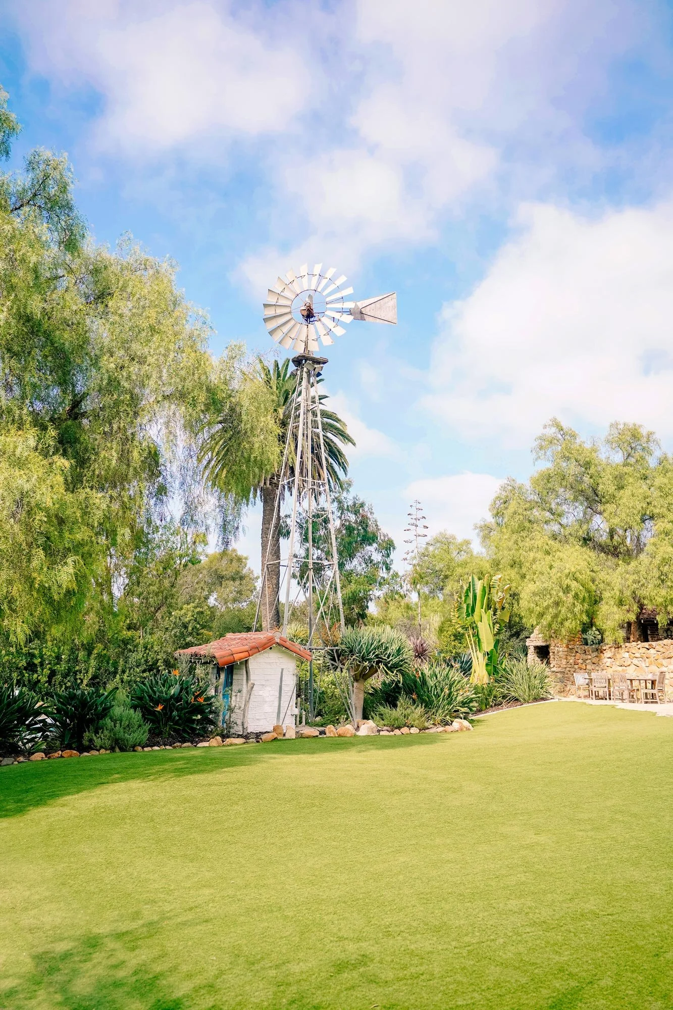 A photo of a large vintage metal windmill standing over a small white shed with a red tiled roof, located in a lush green garden setting with a grassy lawn in the foreground and various trees, including a tall palm tree, under a blue sky with white c