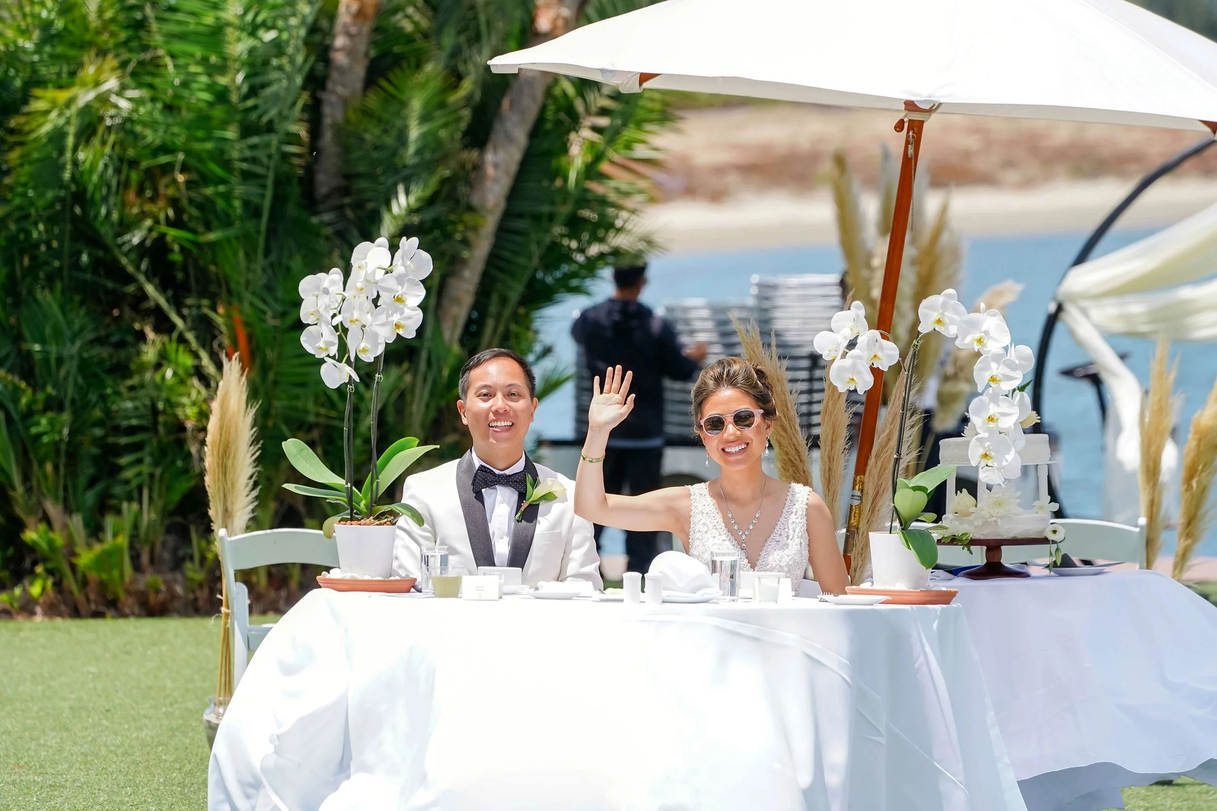 A happy bride and groom are seated at their white-clothed sweetheart table under a large white umbrella during their outdoor wedding reception in Mission Bay, with the bride waving to guests.


