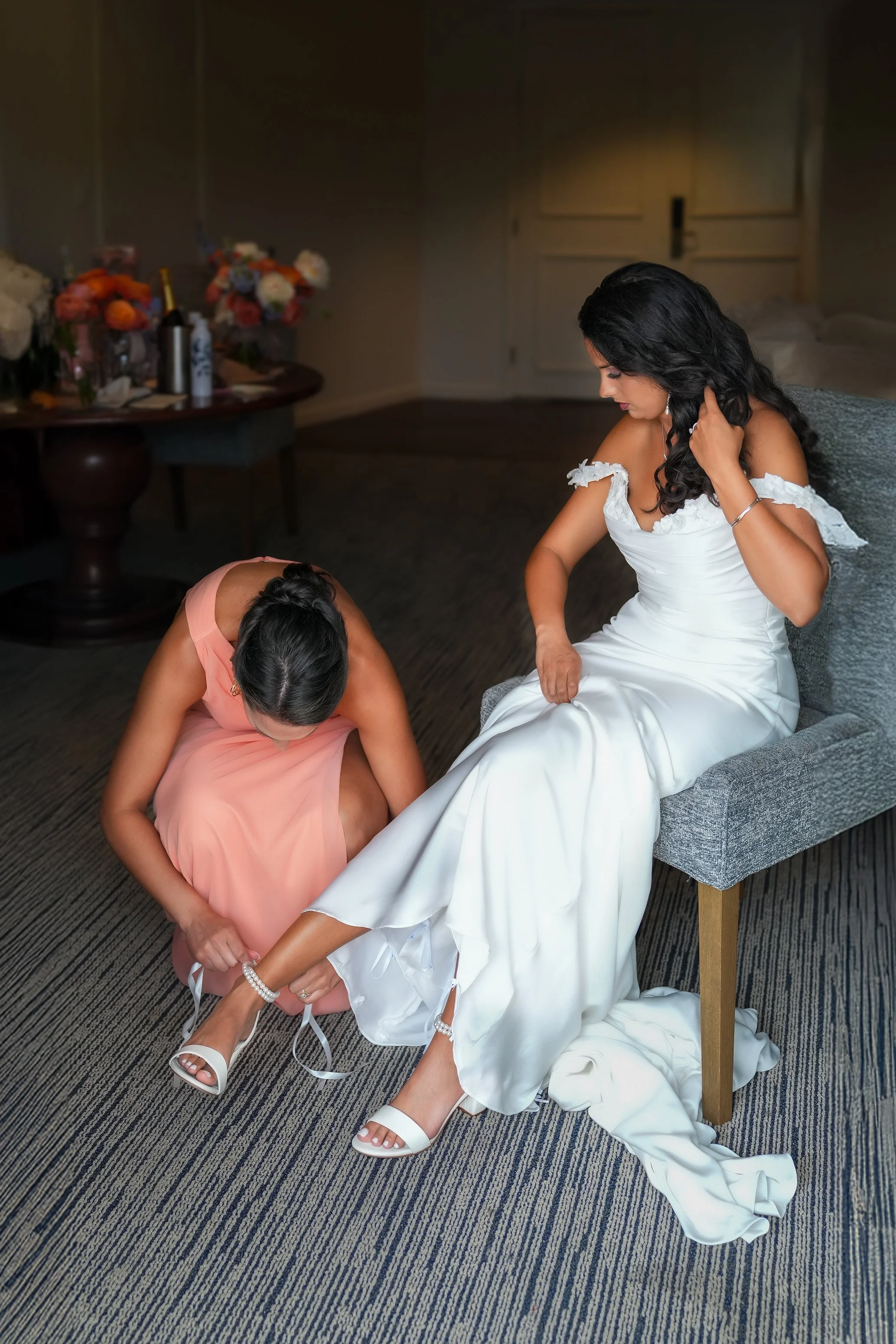 A candid wedding photo by Reimo Photography showing a bridesmaid in a coral dress leaning down to help a bride fasten her white pearl-embellished heels. The bride is seated in a grey chair, wearing a sleek off-the-shoulder white satin wedding gown at