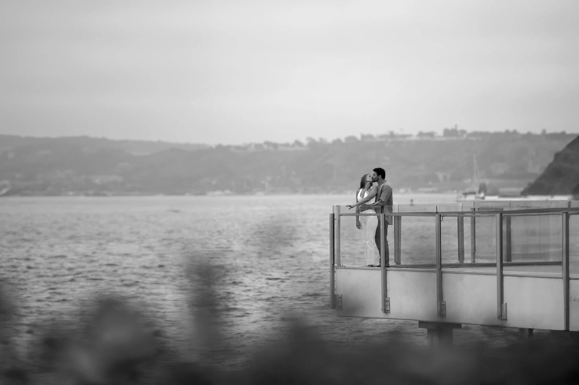 A black and white photograph of a couple embracing on a modern, elevated pier or dock extending over a large body of water. The woman wears a light-colored dress, and the man wears a light shirt and dark trousers. They are standing beside a glass and