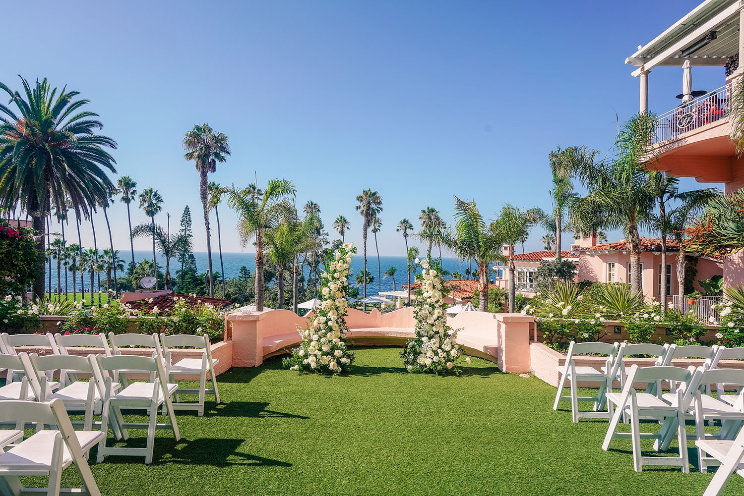 An outdoor wedding ceremony setup at the La Valencia Hotel overlooking the Pacific Ocean, featuring white chairs arranged on green turf, floral arrangements, numerous palm trees, and a clear blue sky.