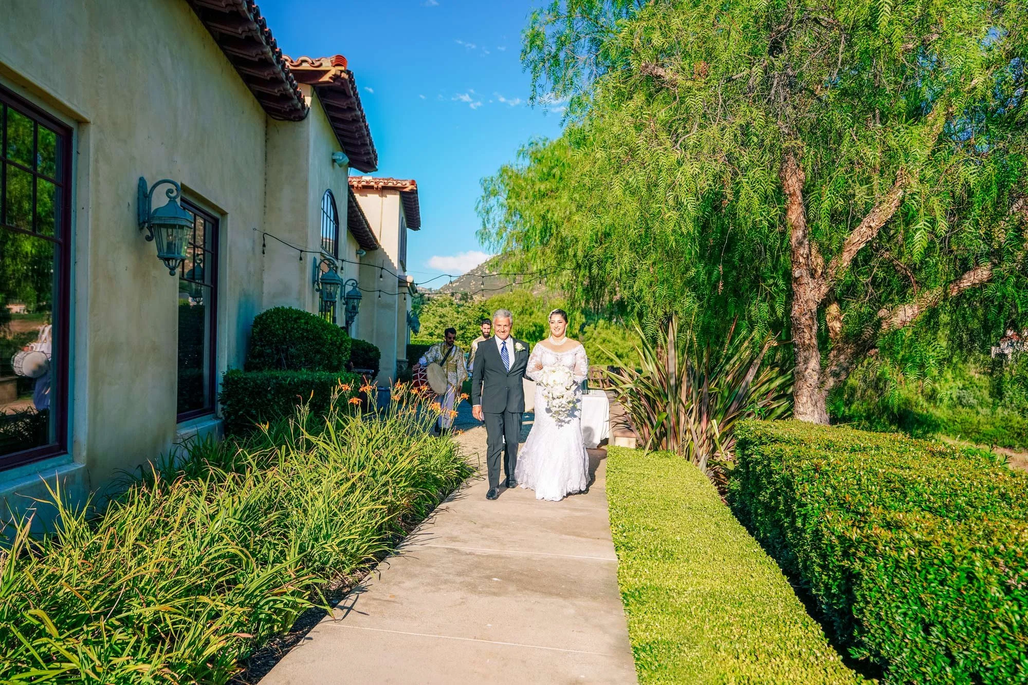A daytime outdoor photograph of a bride in a white long-sleeved lace wedding dress being escorted down a paved walkway by an older man in a dark suit. The walkway is lined with green hedges and plants. They are walking beside a beige stucco building 