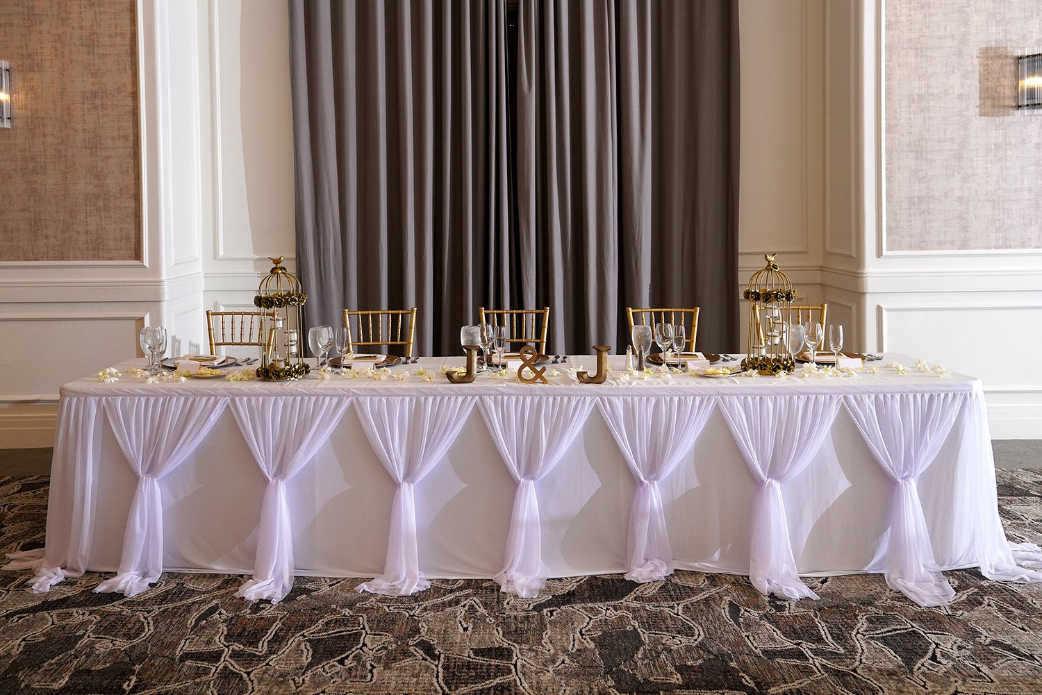 A head table set for an event with a white sheer gathered table skirt, gold chiavari chairs, gold birdcage centerpieces, gold ampersand and "J & J" initials, in front of grey curtains.