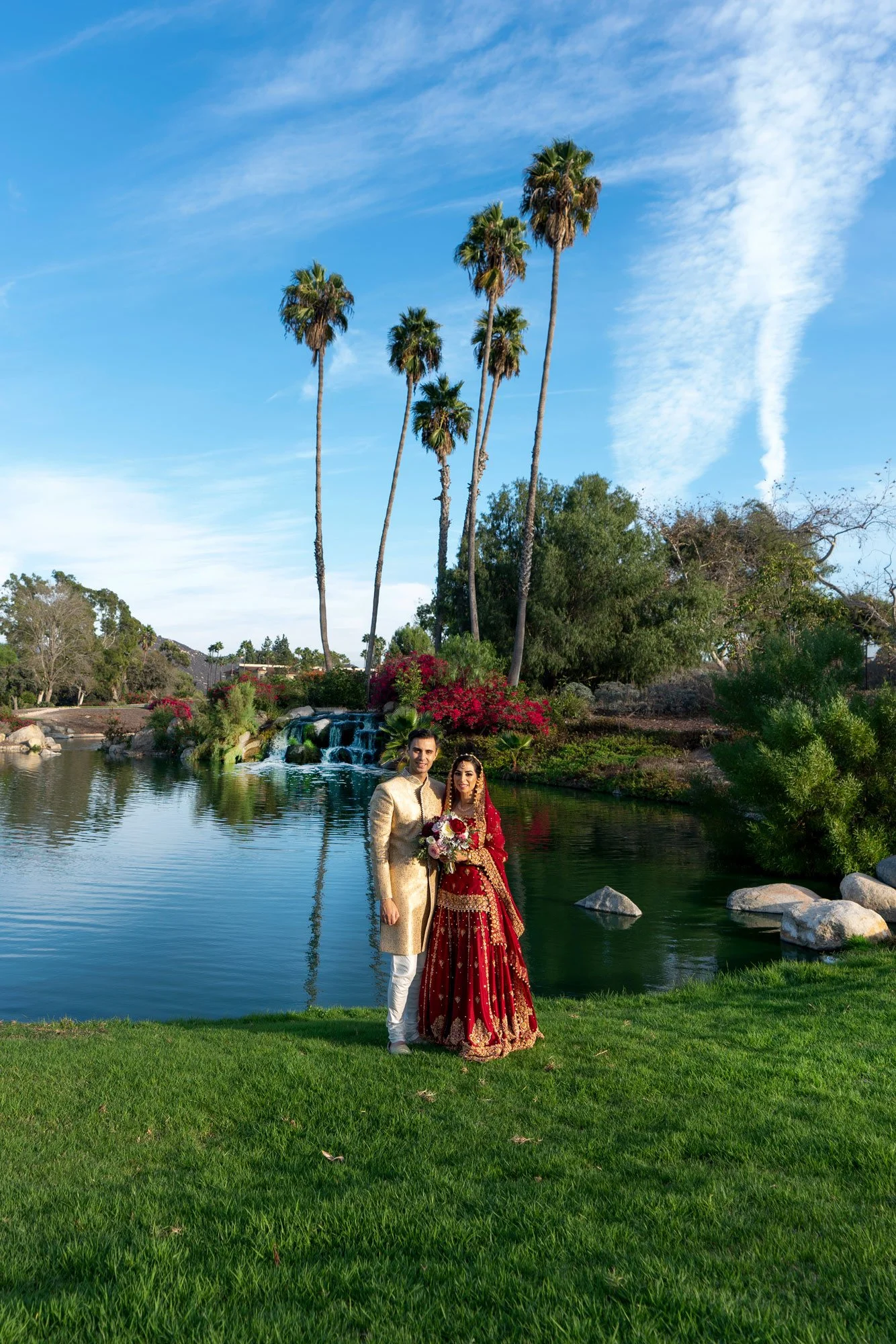 A full-length photograph of a newly married couple posing outdoors in traditional attire. The groom wears a cream-colored outfit with gold accents, and the bride wears an ornate, deep red and gold lehenga. They stand on a grassy lawn beside a calm po