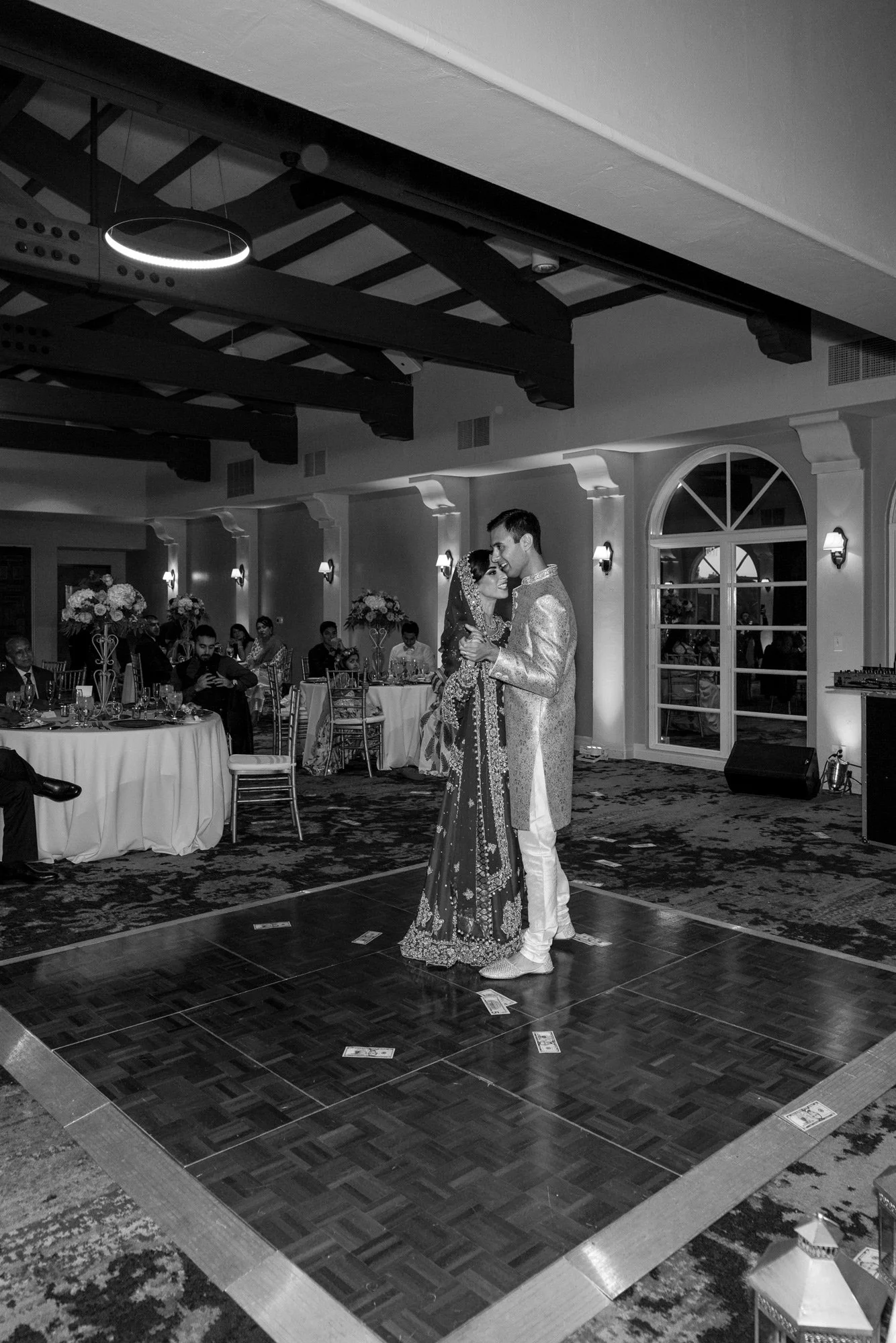 A black and white photograph of a newly married couple dancing together on a checkered wooden dance floor. The bride wears an ornate, dark, traditional outfit, and the groom wears a light-colored suit or sherwani. Guests are seated at tables in the b