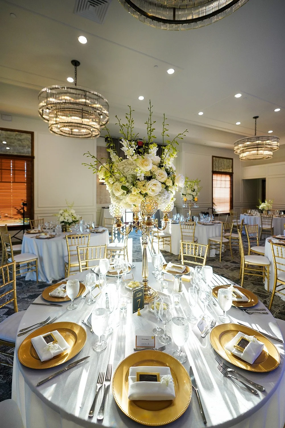 An elegant indoor dining table set for a formal event, featuring gold charger plates, white napkins, silver cutlery, gold chiavari chairs, and a tall white and green floral centerpiece.
