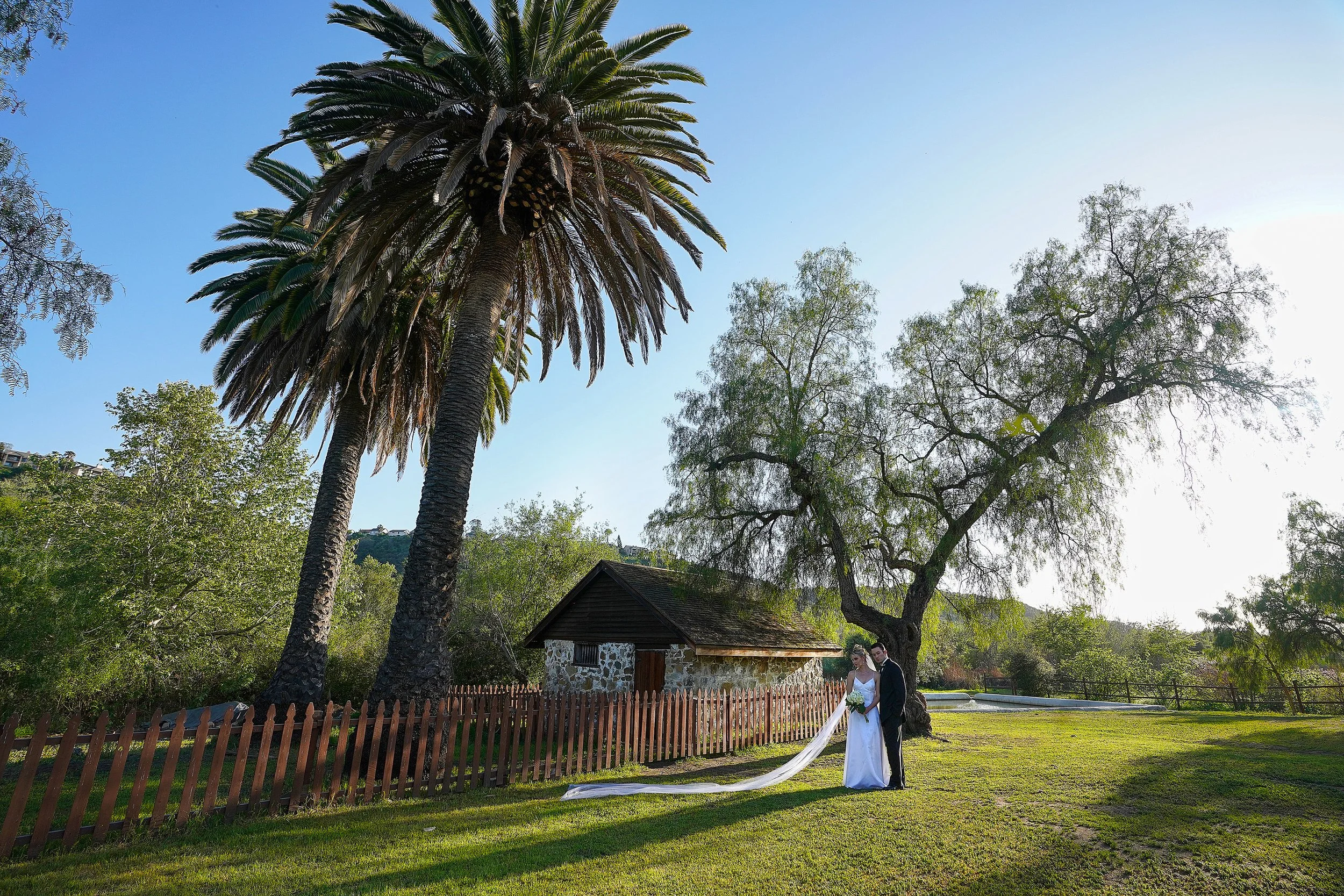 A bride and groom pose for a photo in front of a historic stone spring house with tall palm trees in Rancho Penasquitos Ranch House.
