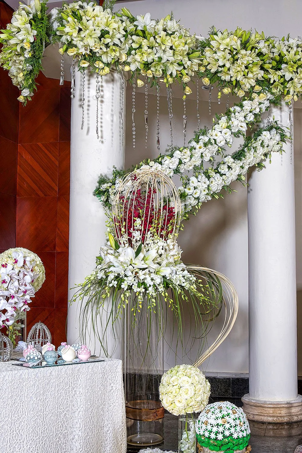 An elaborate, formal indoor floral display for an event, featuring a large white and green floral arch with hanging crystal accents, white columns, and a tall glass vase centerpiece with a red and white flower arrangement inside a wire birdcage.