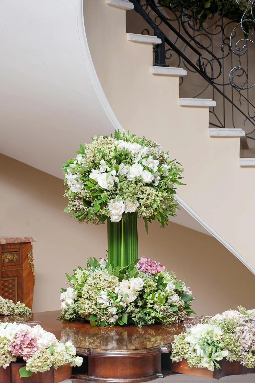 A large, elegant white and green floral arrangement in a tall glass vase on a wooden table in an entryway, with a curved staircase and wrought-iron railing in the background.