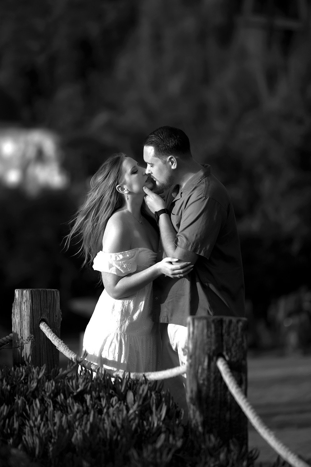 A black and white outdoor photograph of a couple embracing and about to kiss. The woman wears an off-the-shoulder white dress, and the man wears a dark shirt and light-colored pants. They stand near a rope and wooden post barrier in the foreground, a