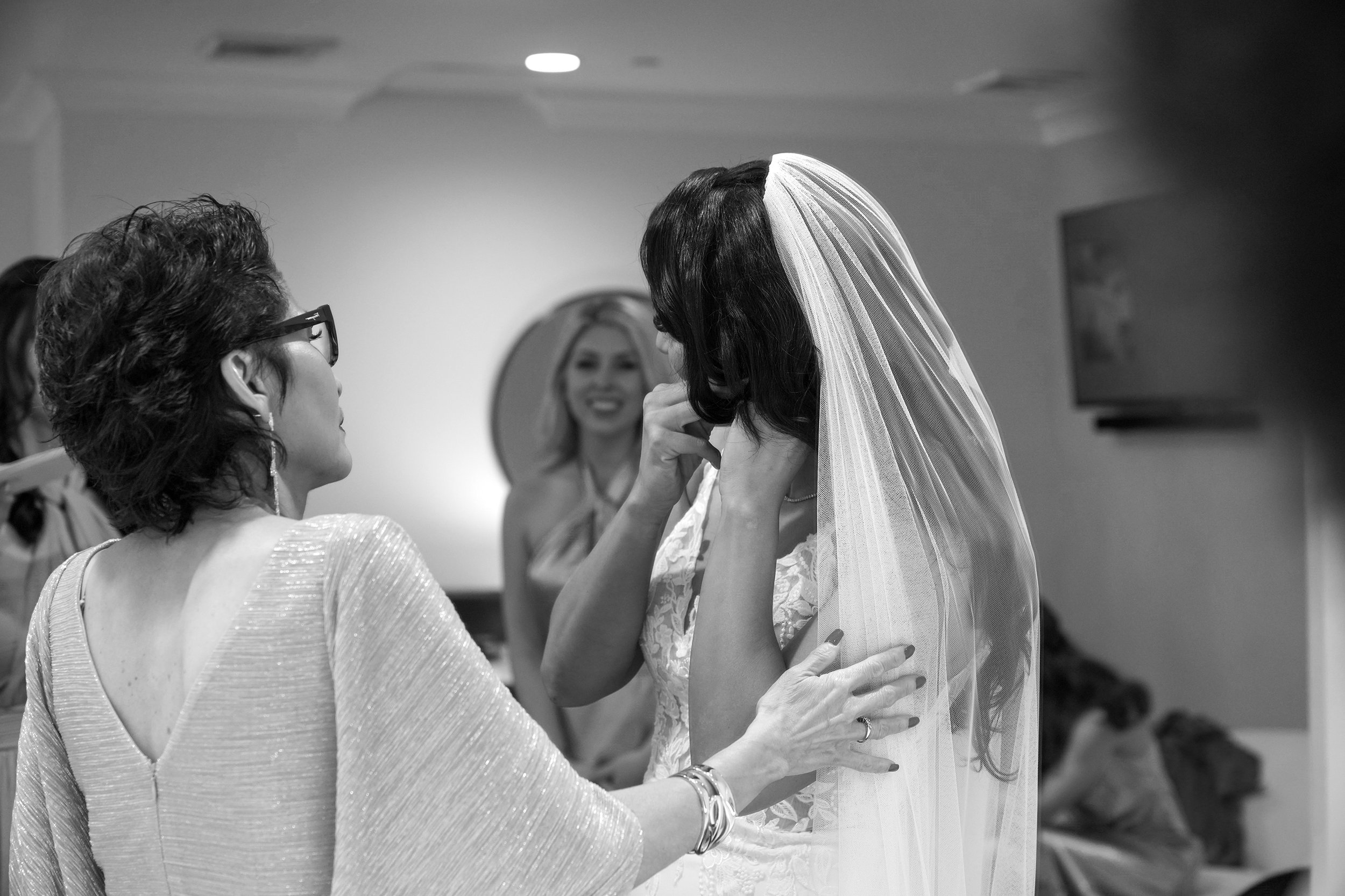 In this intimate black and white photograph, a bride prepares for her wedding day with the help of an older woman, likely a mother or close relative. The bride, seen in profile with her long, dark hair partially covered by a floor-length sheer veil, 