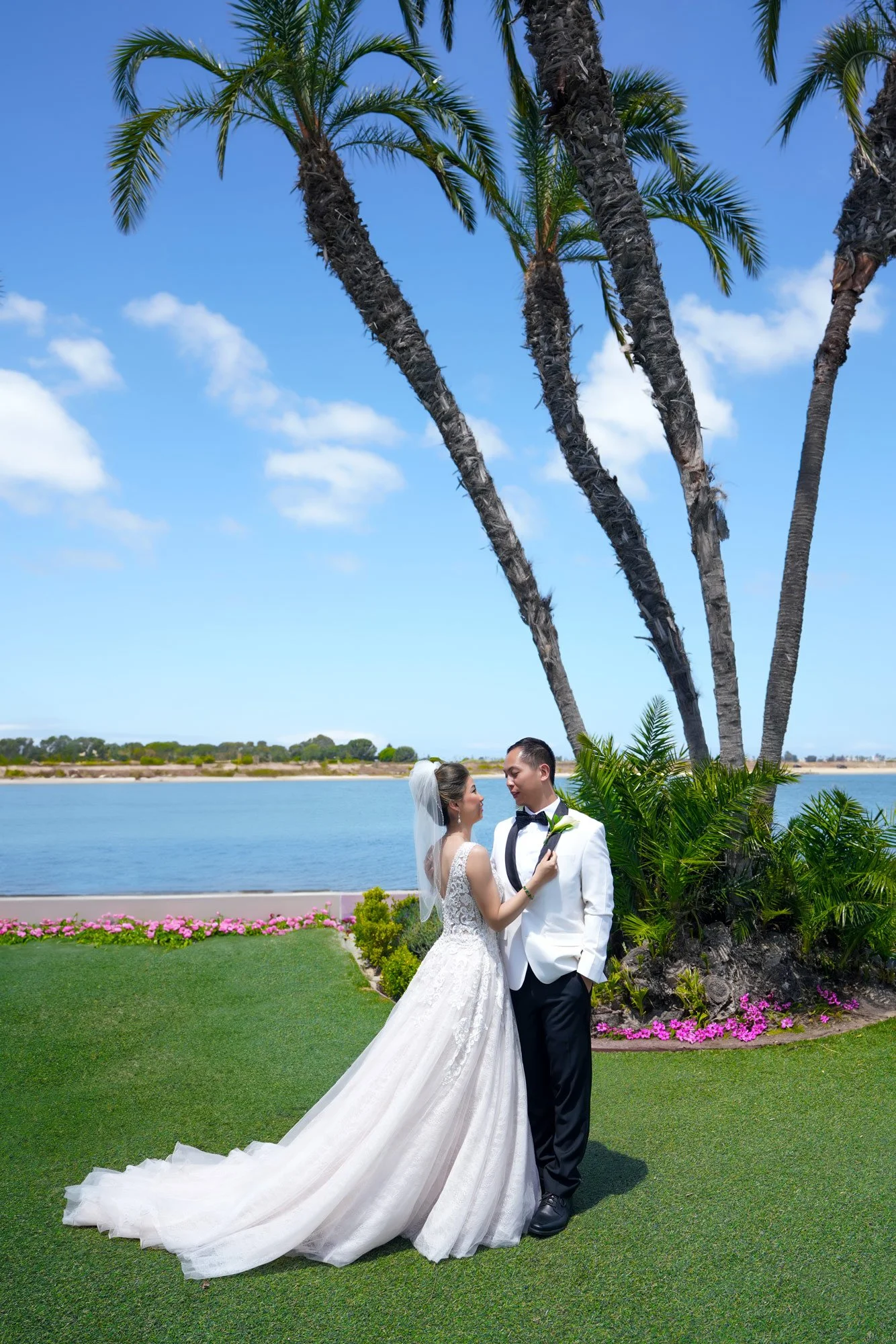 A bride in a white lace gown and a groom in a white tuxedo jacket stand facing each other on a green lawn with a water view of Mission Bay and tall palm trees in the background.