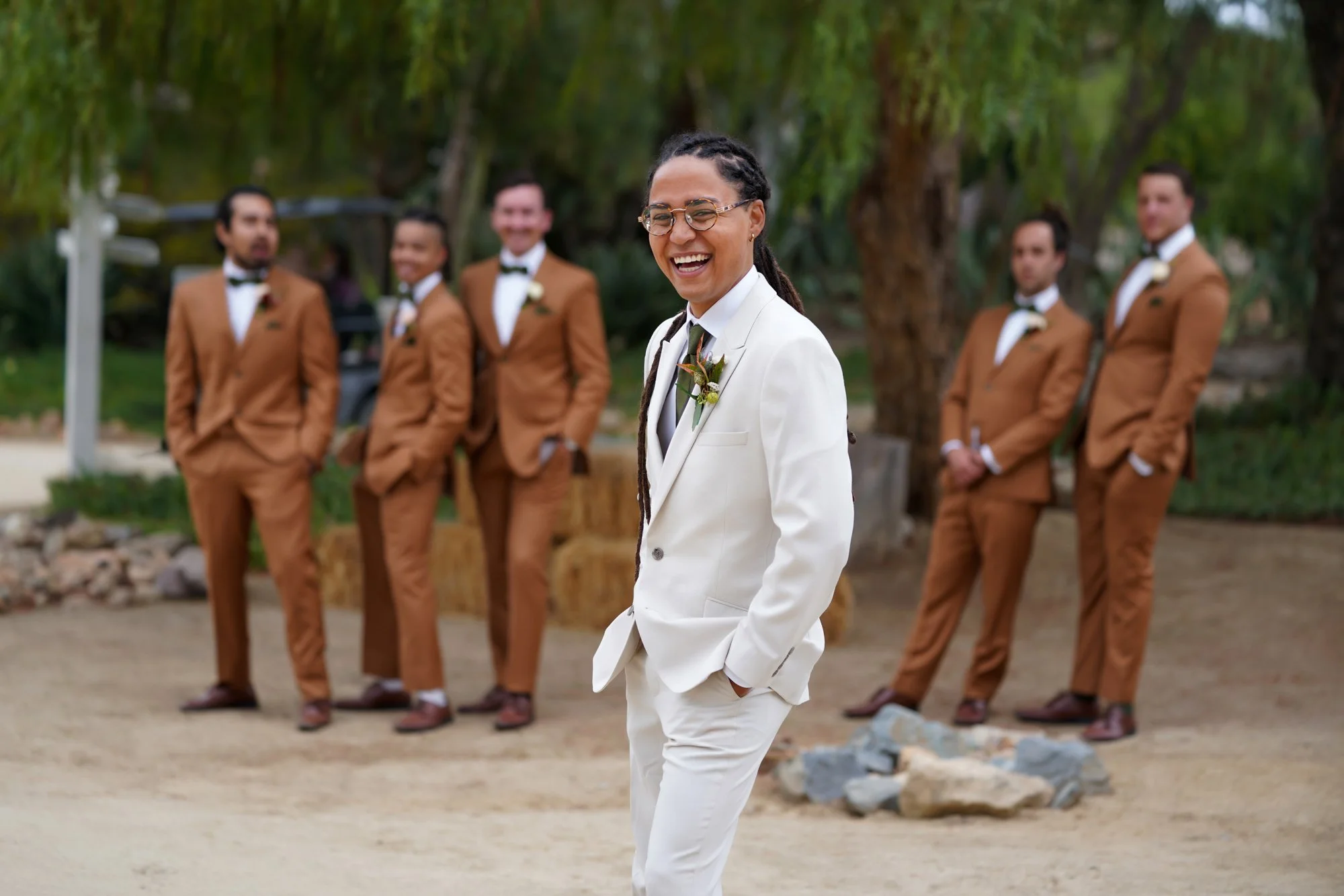 A photo of a groom in a cream-colored suit and glasses laughing and looking at the camera, with five other people in brown suits standing in the background outdoors on a dirt path.