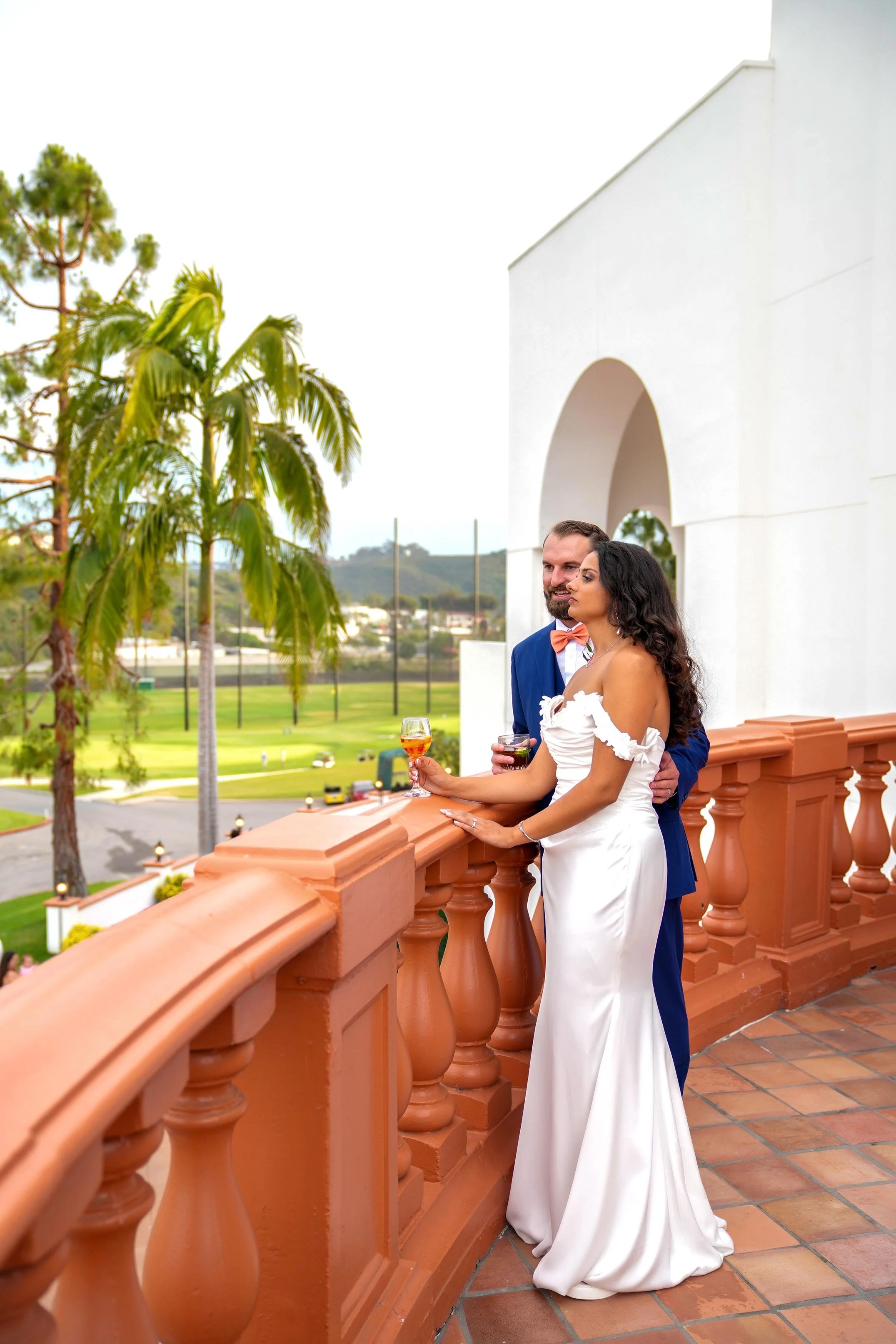 A bride in a white off-the-shoulder gown and a groom in a blue suit with an orange bowtie stand on a terracotta-railed balcony, each holding a drink, and look out over La Costa golf course and distant hills with palm trees.