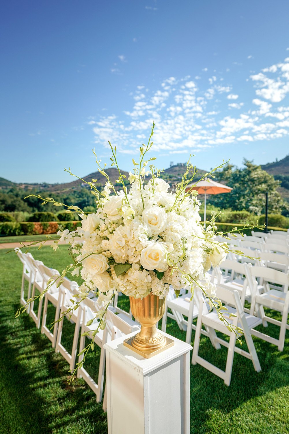 A large white floral arrangement in a gold urn-style vase, placed on a white pedestal on a grassy lawn. Rows of white folding chairs are set up in the background for a wedding ceremony, with green hills visible in the distance under a blue sky with w