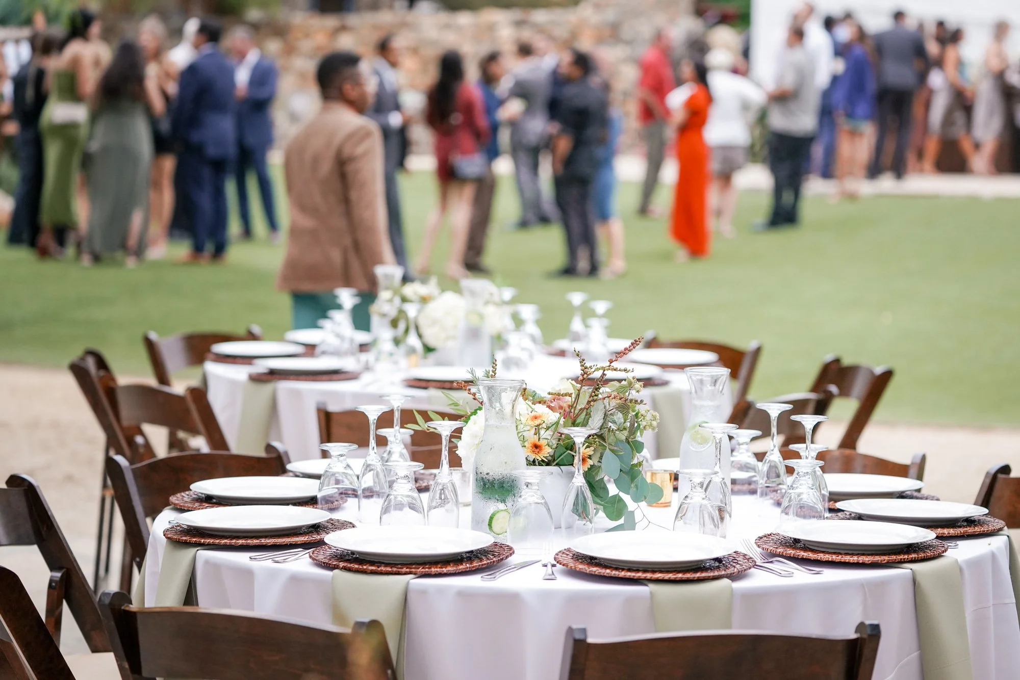 A photo of an outdoor event reception venue featuring dining tables set with white linens and dark wooden chairs in the foreground, and a grassy area with many formally dressed guests mingling in the background.


