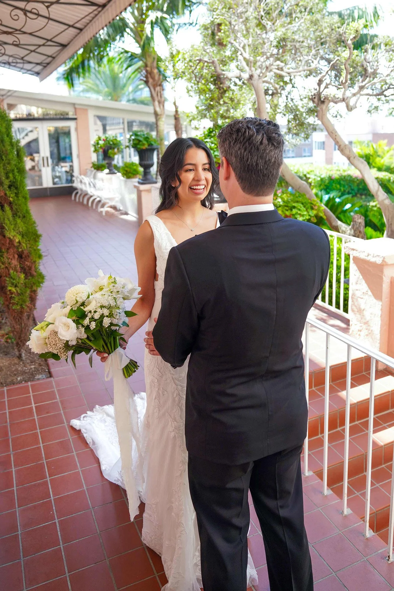 An outdoor photograph of a smiling bride in a white lace wedding dress holding a white floral bouquet, looking at the groom, who is wearing a black tuxedo and standing with his back to the camera. They are on a red-tiled patio with white railings, an