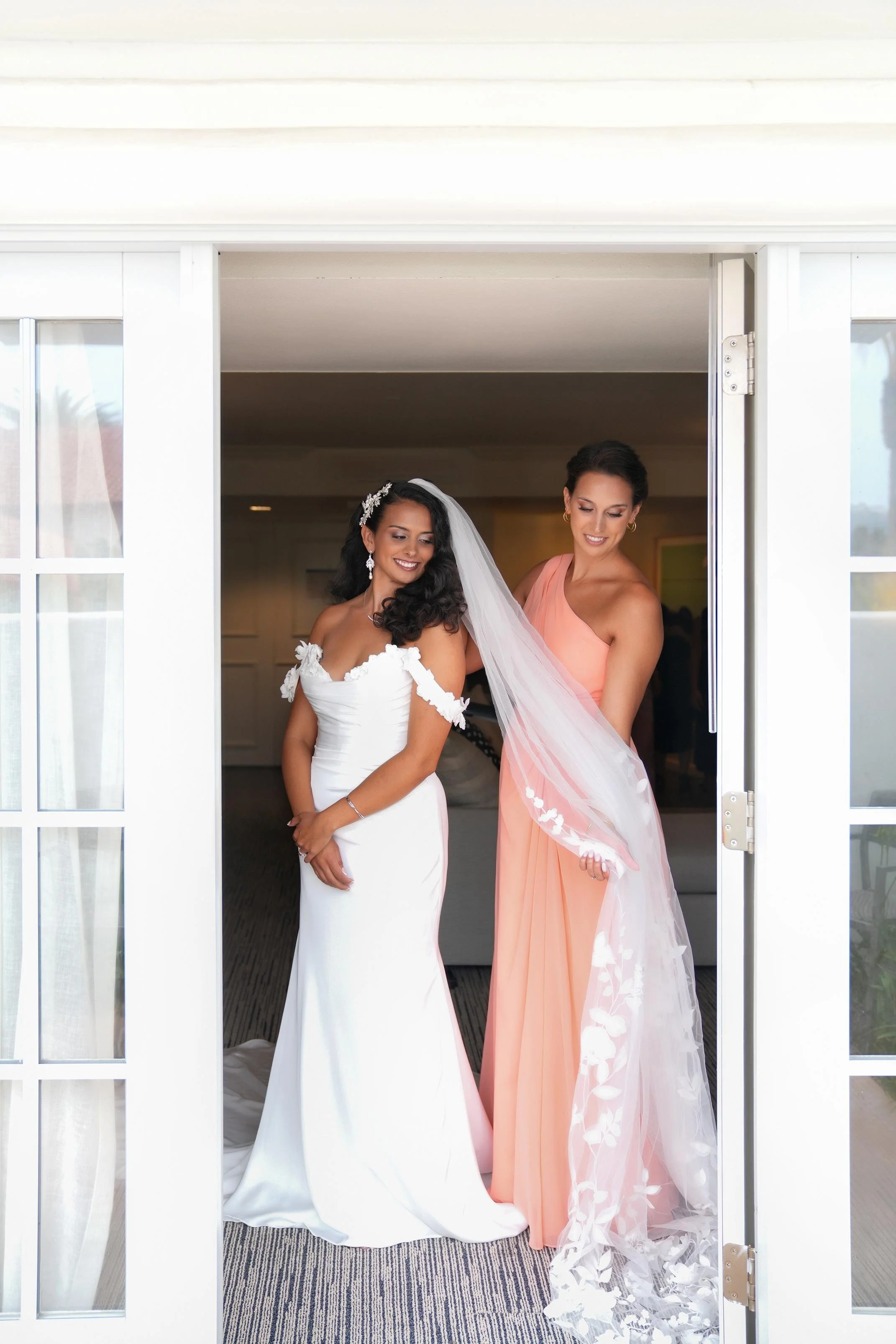 A bride in a white, off-the-shoulder gown and a bridesmaid in a peach dress stand in a doorway, smiling at the camera as the bridesmaid helps hold the bride's long, lace-trimmed veil.


