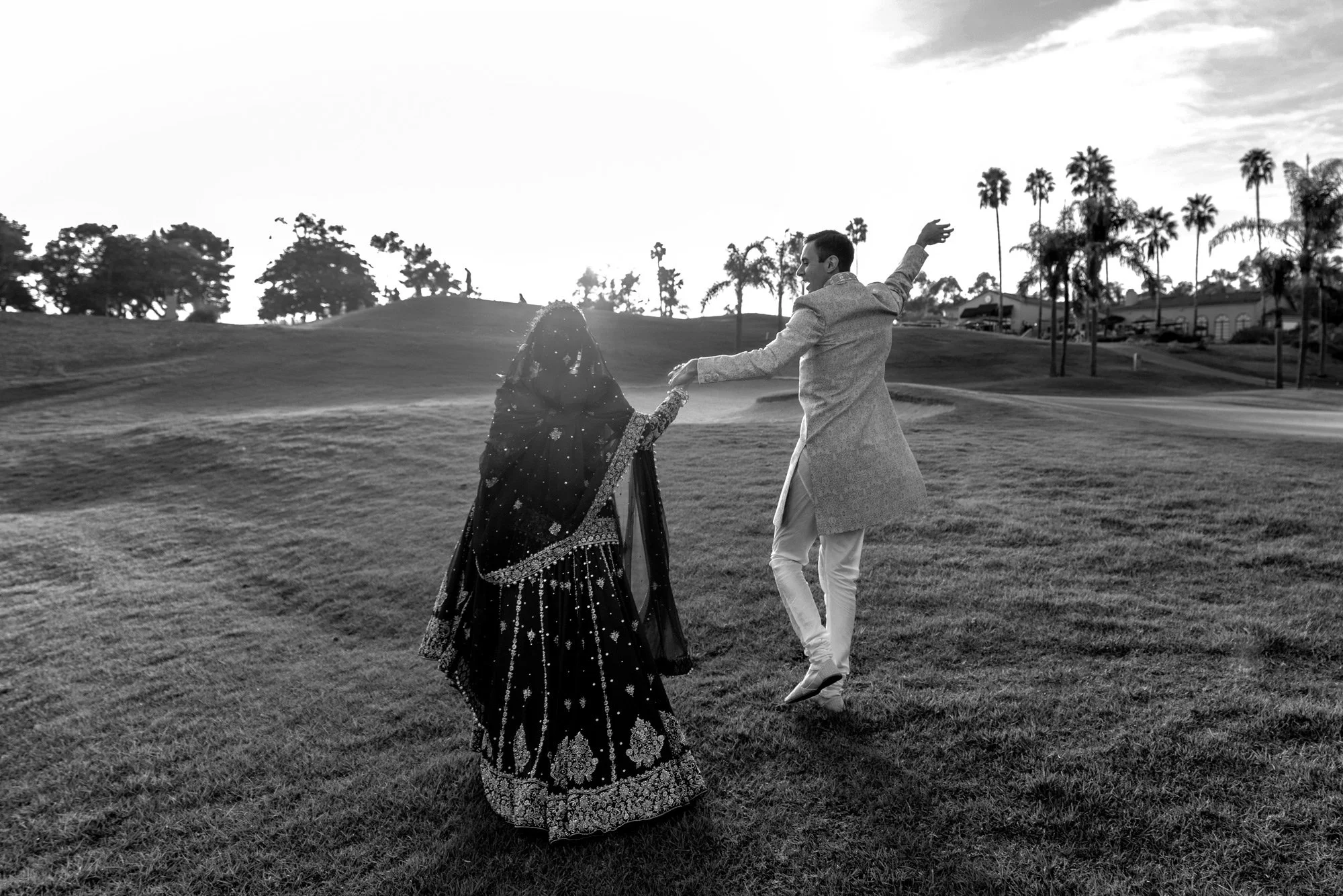 A black and white photograph of a couple in traditional Indian wedding attire running or dancing in a grassy field at sunset. The person on the left is wearing a dark, heavily embroidered lehenga and veil, seen from behind. The person on the right, p