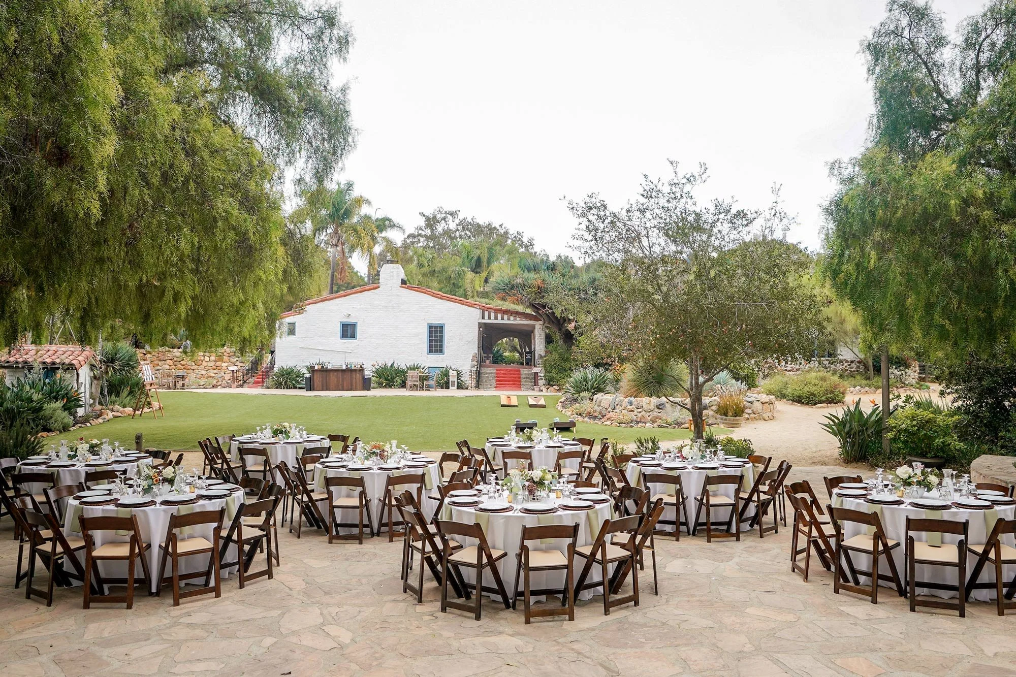 A photo of an outdoor event venue featuring multiple dining tables with white linens and dark wooden chairs on a flagstone patio, with a grassy area and a white hacienda-style building in the background, surrounded by lush greenery in Carlsbad.