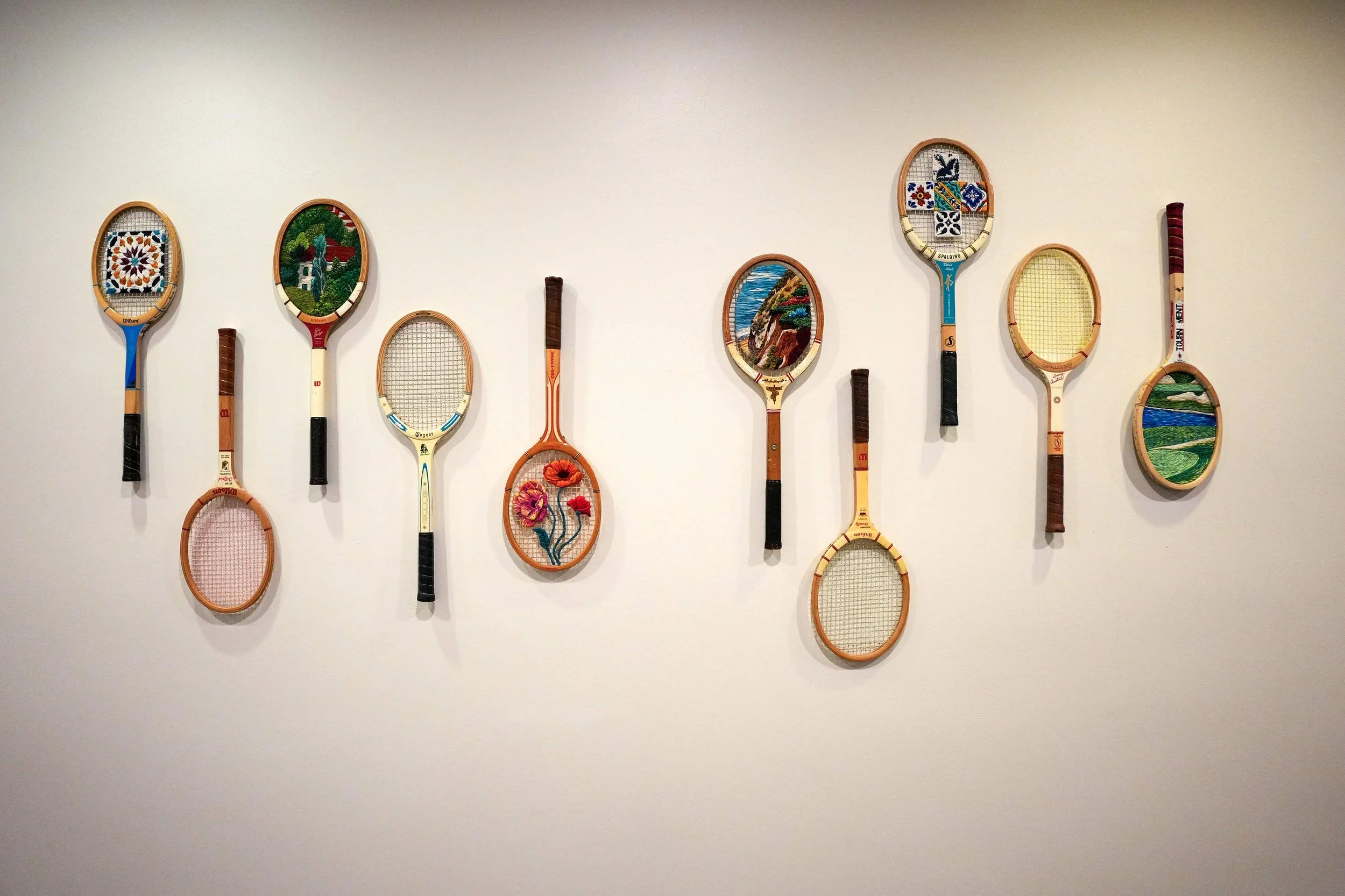 A collection of vintage wooden tennis rackets hanging on a white wall, with the string beds replaced by circular embroidered artworks featuring various designs, including mosaic tiles, landscapes, and flowers.