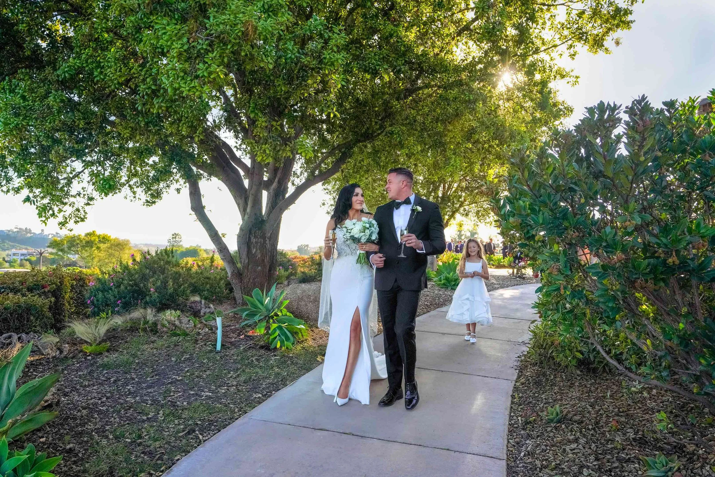 An outdoor photograph of a newly married couple walking down a paved walkway after their wedding ceremony. The bride wears a long white dress with a high leg slit and holds a white floral bouquet, and the groom wears a black tuxedo and holds a champa