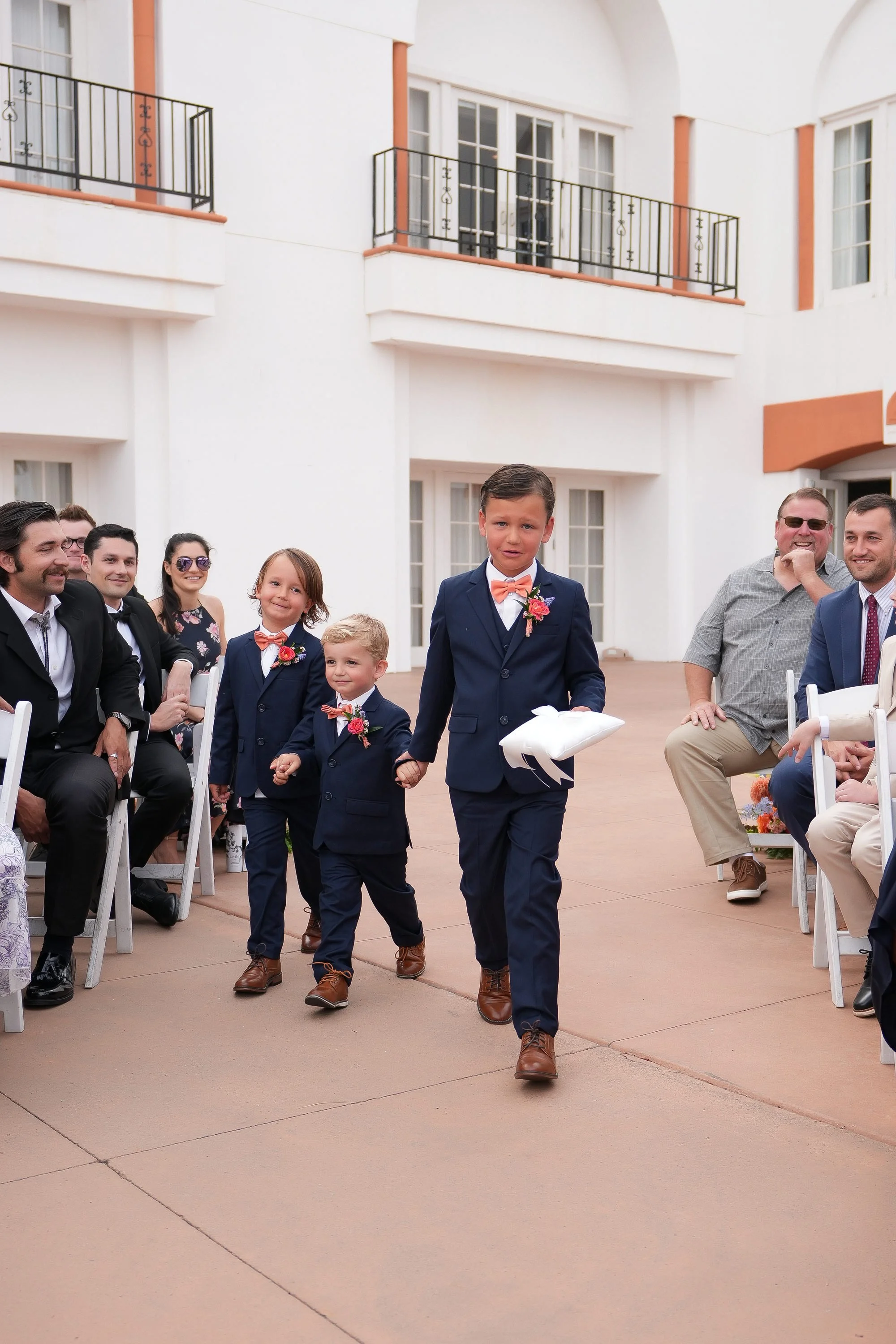 Two young boys dressed in matching navy blue suits and orange bowties walk down an outdoor aisle holding hands, followed by an older boy in the same attire carrying a white object, as wedding guests watch from the sides.