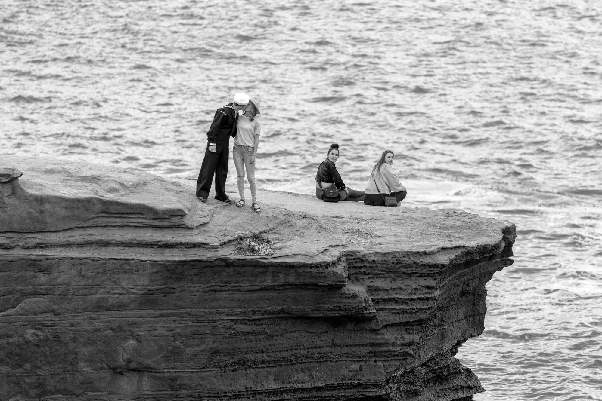 A black and white photograph capturing people on a rocky cliff, overlooking the ocean. On the left, a man in a sailor's uniform is embracing a woman. To their right, two other women are seated on the edge of the cliff, looking out at the wavy water.
