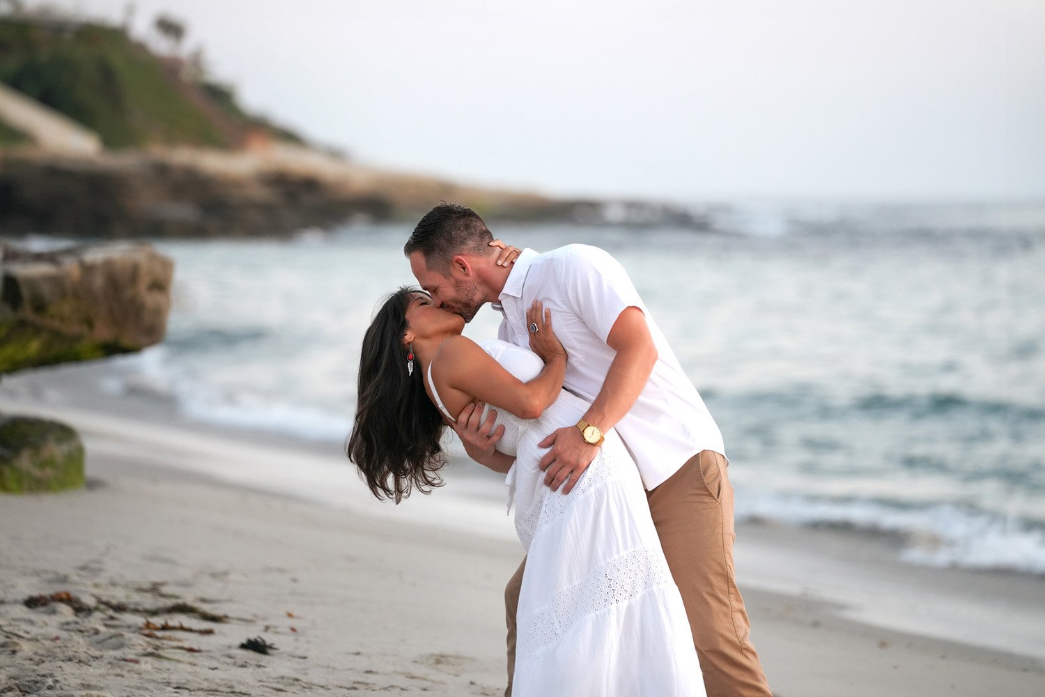 A couple embracing and kissing on a sandy beach. The woman wears a white dress, and the man wears a white short-sleeved shirt and tan pants. Waves are rolling in on the shore, and a rocky bluff is visible in the background on the left.