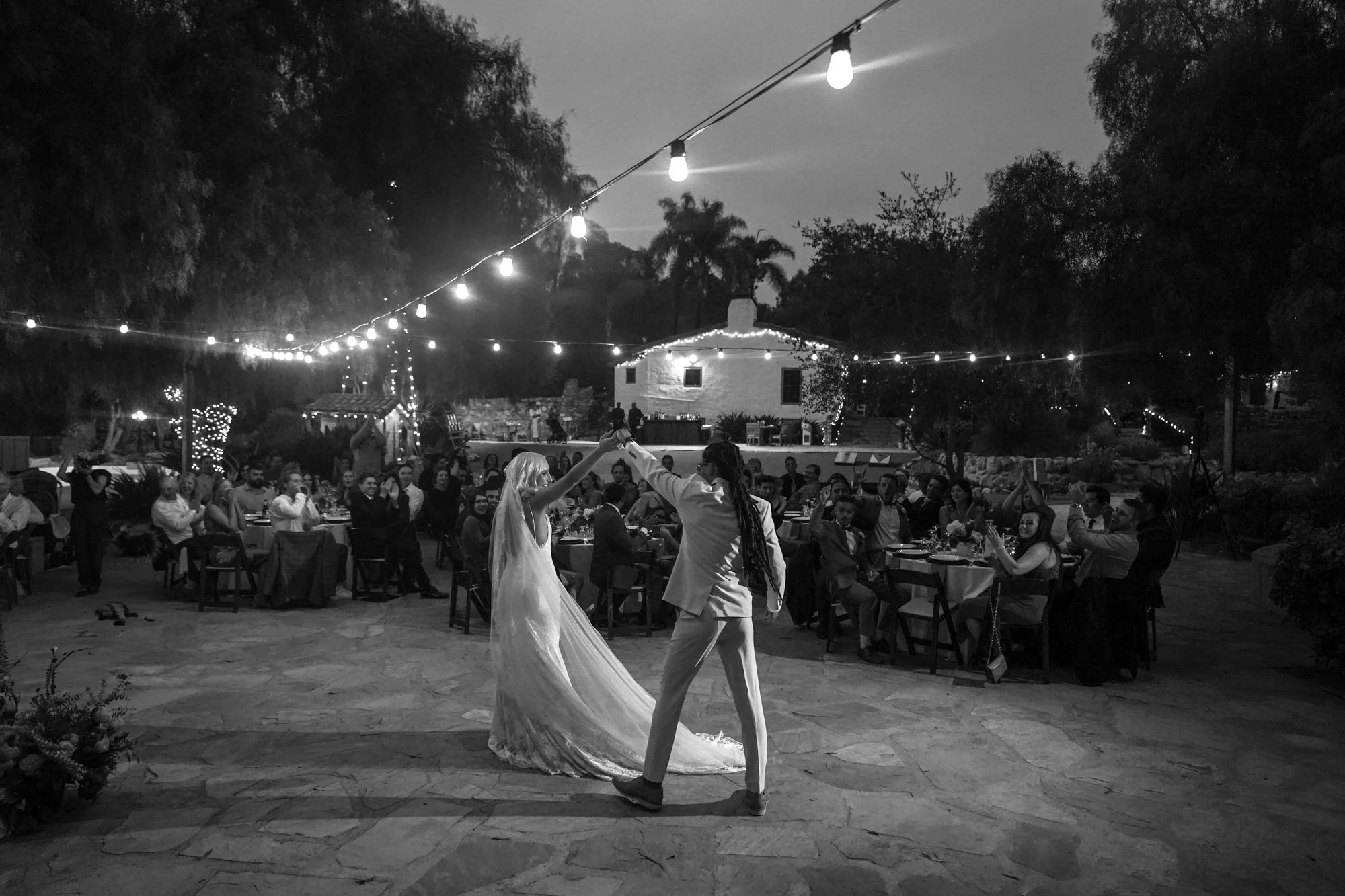 A black and white photo of a newly married couple dancing together on an outdoor flagstone patio during their wedding reception. String lights hang overhead, illuminating the couple and seated guests at round tables, with a white hacienda-style build
