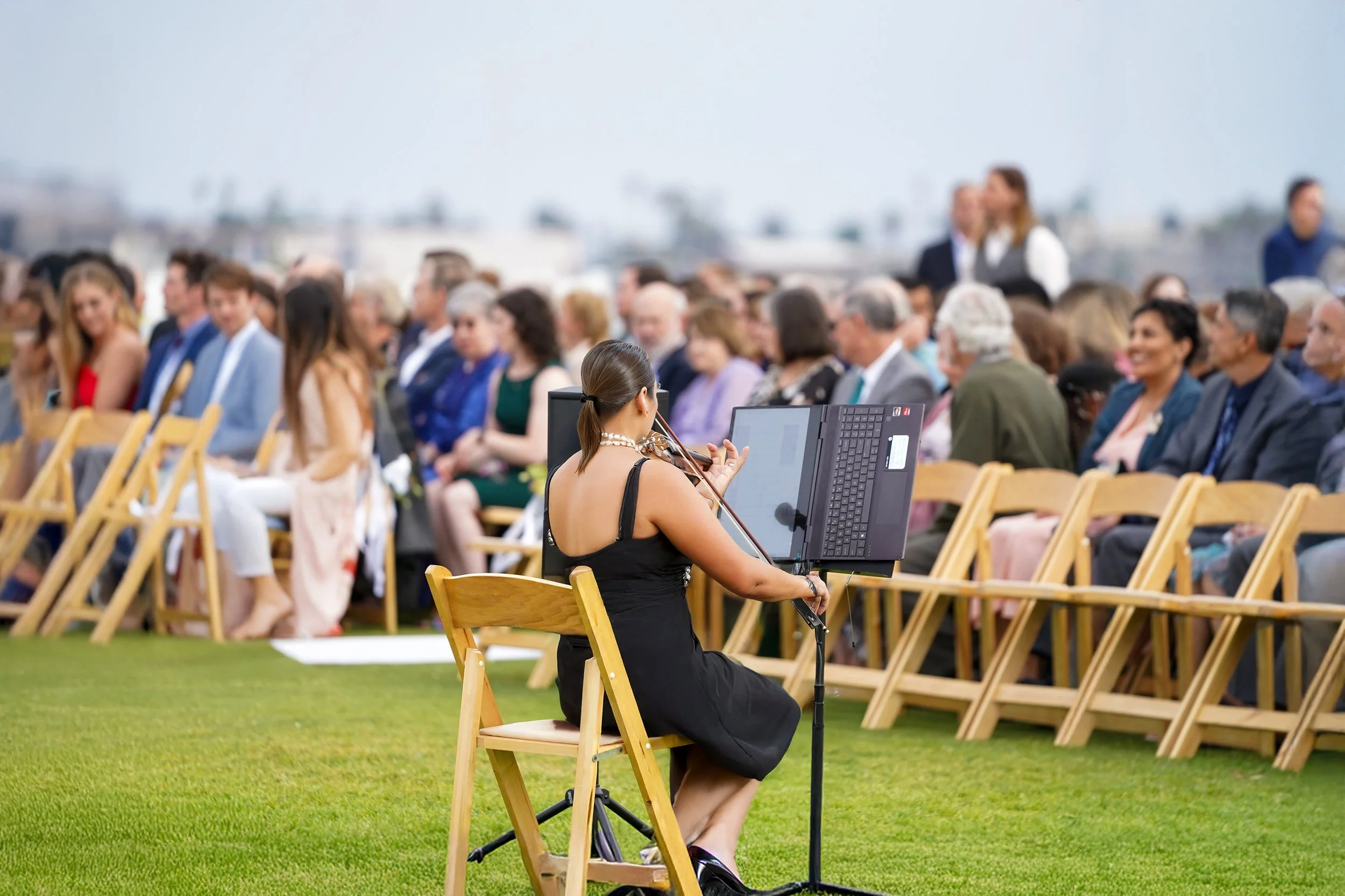 A female violinist in a black dress performs on a grassy lawn during an outdoor wedding ceremony with guests seated in wooden chairs.