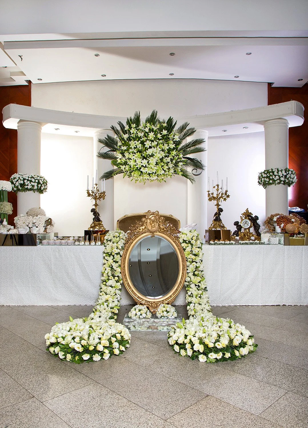 An elaborate, formal indoor event setup, possibly a Persian wedding Sofreh Aghd, featuring a large ornate gold mirror, candelabras, extensive white floral arrangements draped around the mirror and on the floor, and white tablecloths in a venue with l