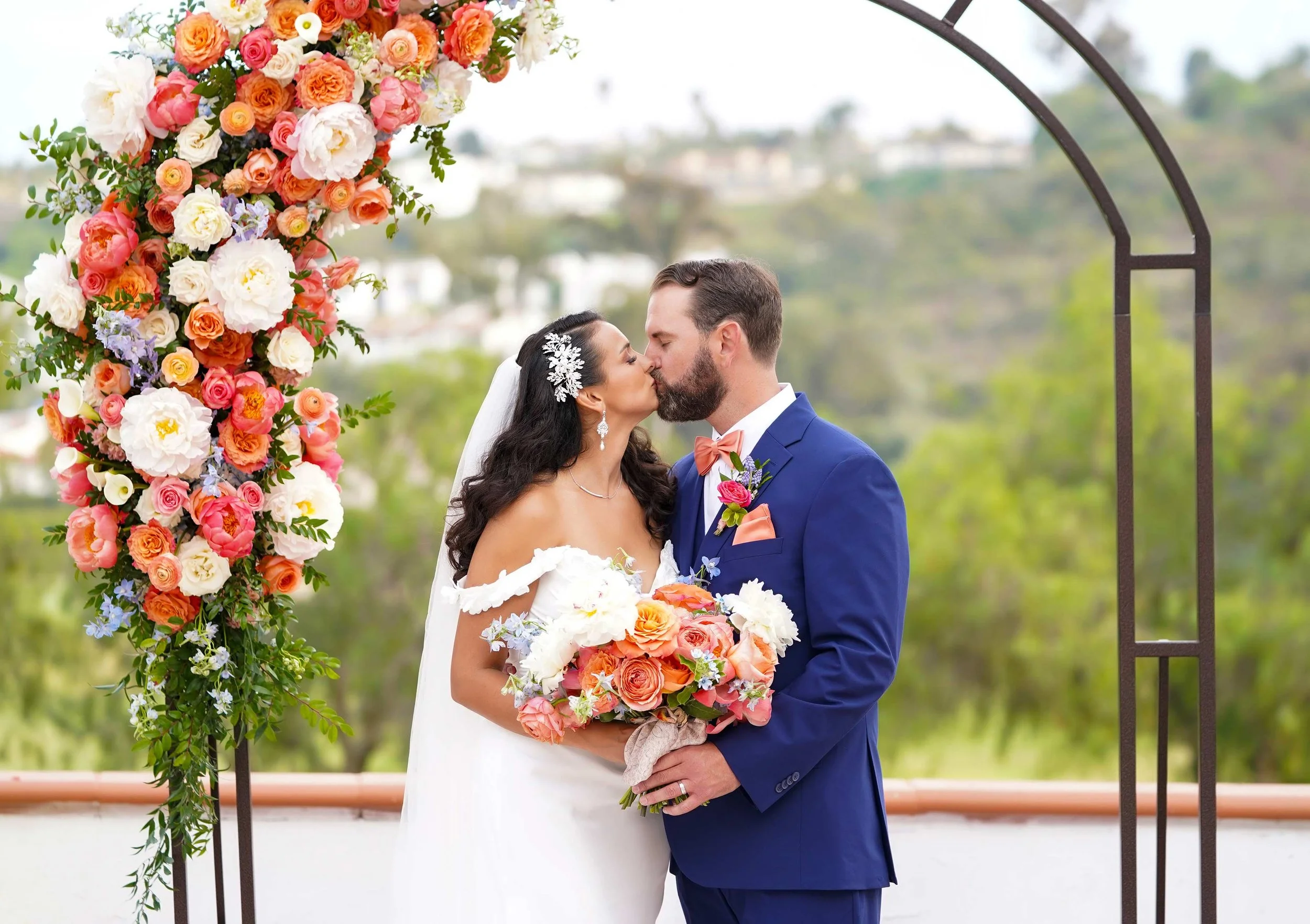 A bride in a white, off-the-shoulder gown and a groom in a blue suit with an orange bowtie share a kiss under a dark metal arch adorned with large orange, pink, and white floral arrangements in San Diego.


