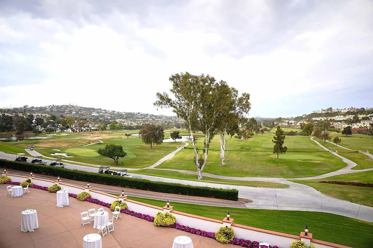 An elevated view of an outdoor event patio set up with white tables and chairs, overlooking a green golf course with trees, cart paths, a small pond, and a residential hillside in the background under an overcast sky.