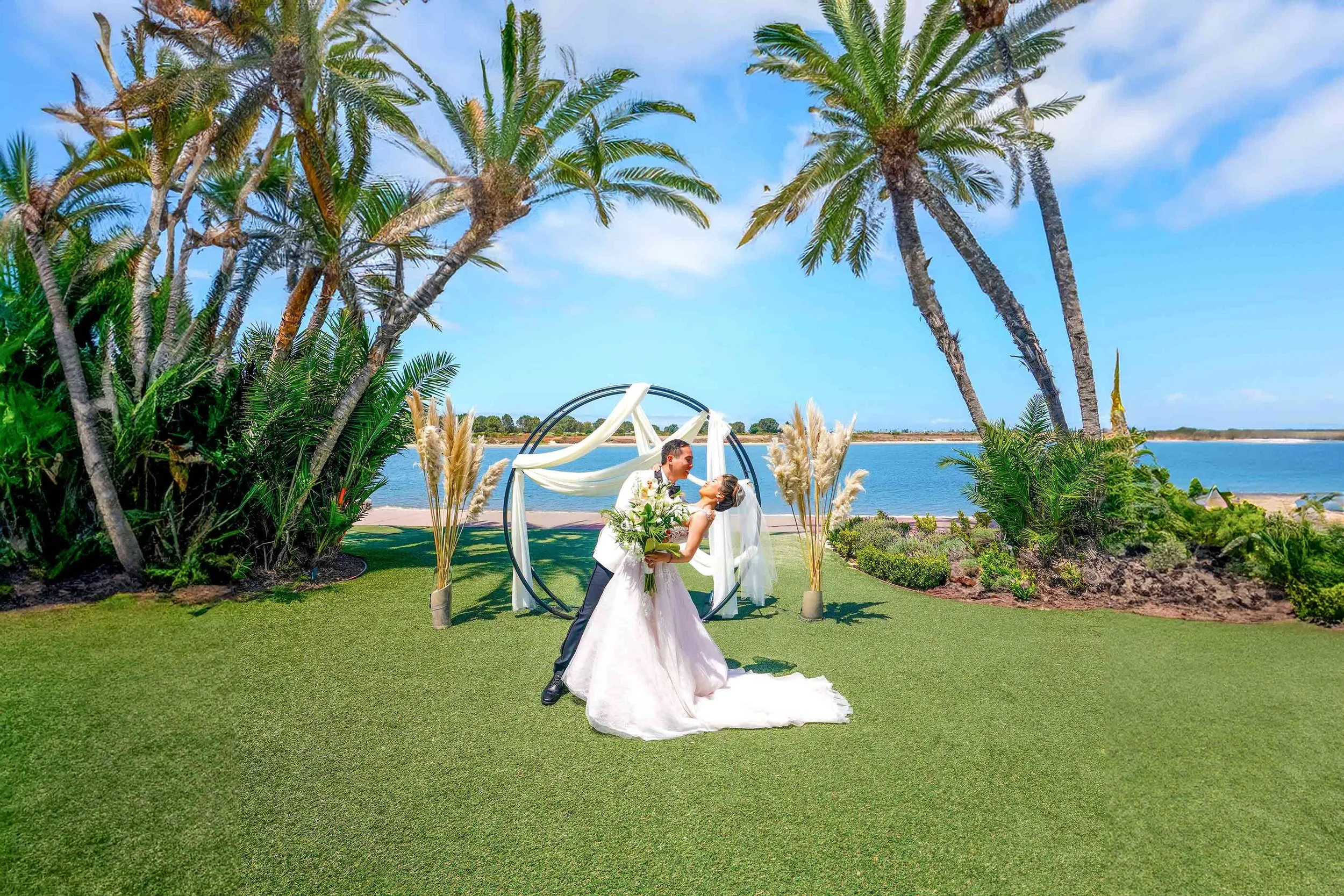 A bride and groom kiss under a modern circular archway decorated with white drapes and pampas grass, surrounded by lush greenery and palm trees overlooking the water during their sunny outdoor wedding ceremony in San Diego.



