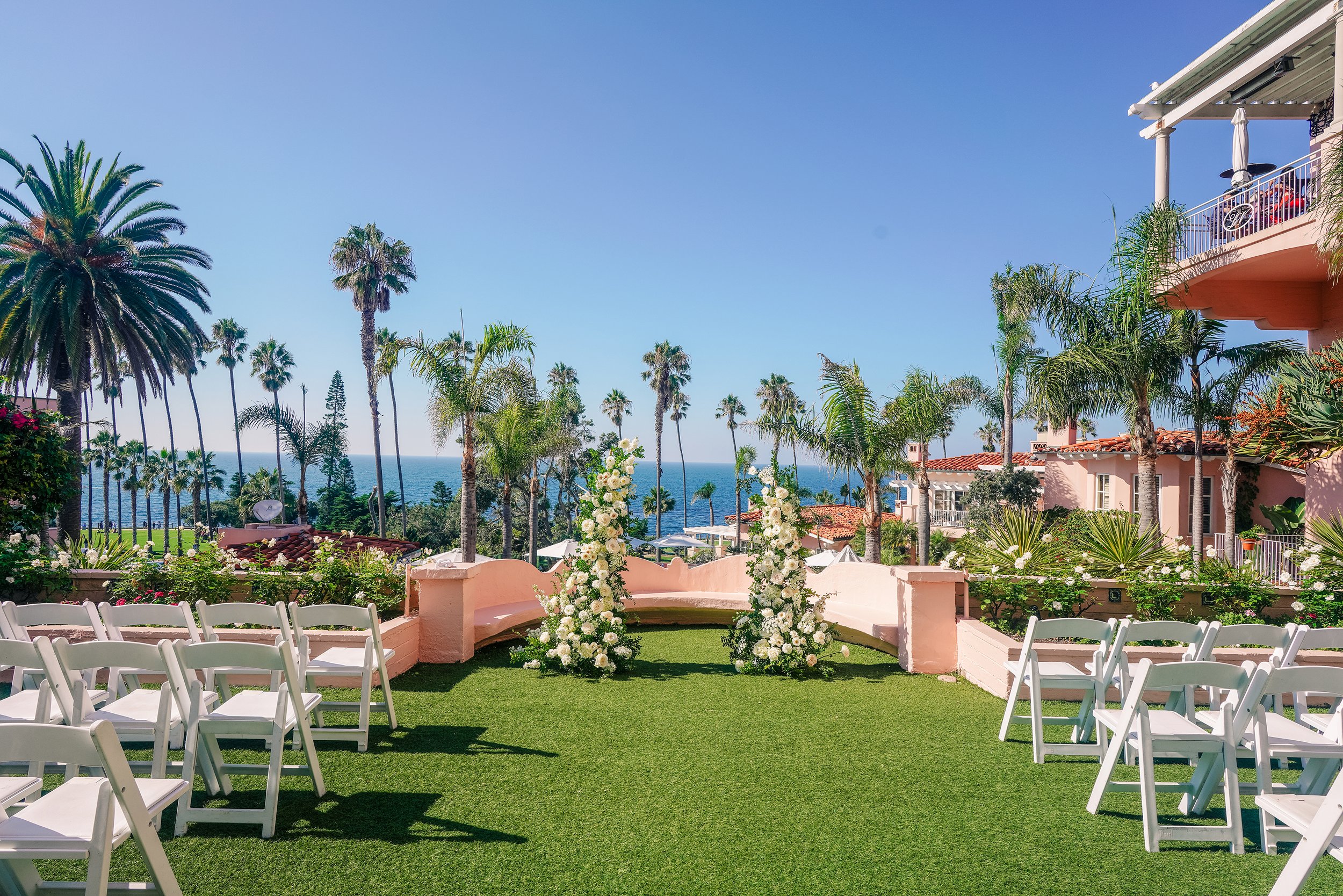An outdoor wedding ceremony setup at the La Valencia Hotel overlooking the Pacific Ocean, featuring white chairs arranged on green turf, floral arrangements, numerous palm trees, and a clear blue sky.