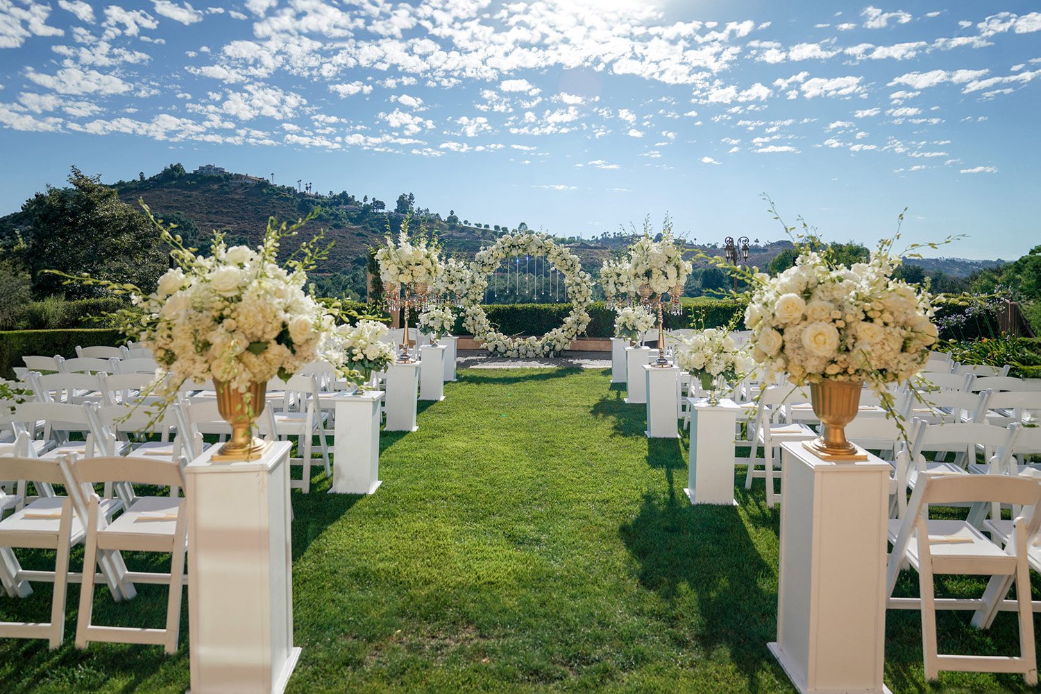 White folding chairs line the aisle, which is marked by white pedestals topped with gold urns holding elaborate white floral arrangements, leading towards a large circular white floral arch. Rolling green hills are visible in the background under a b
