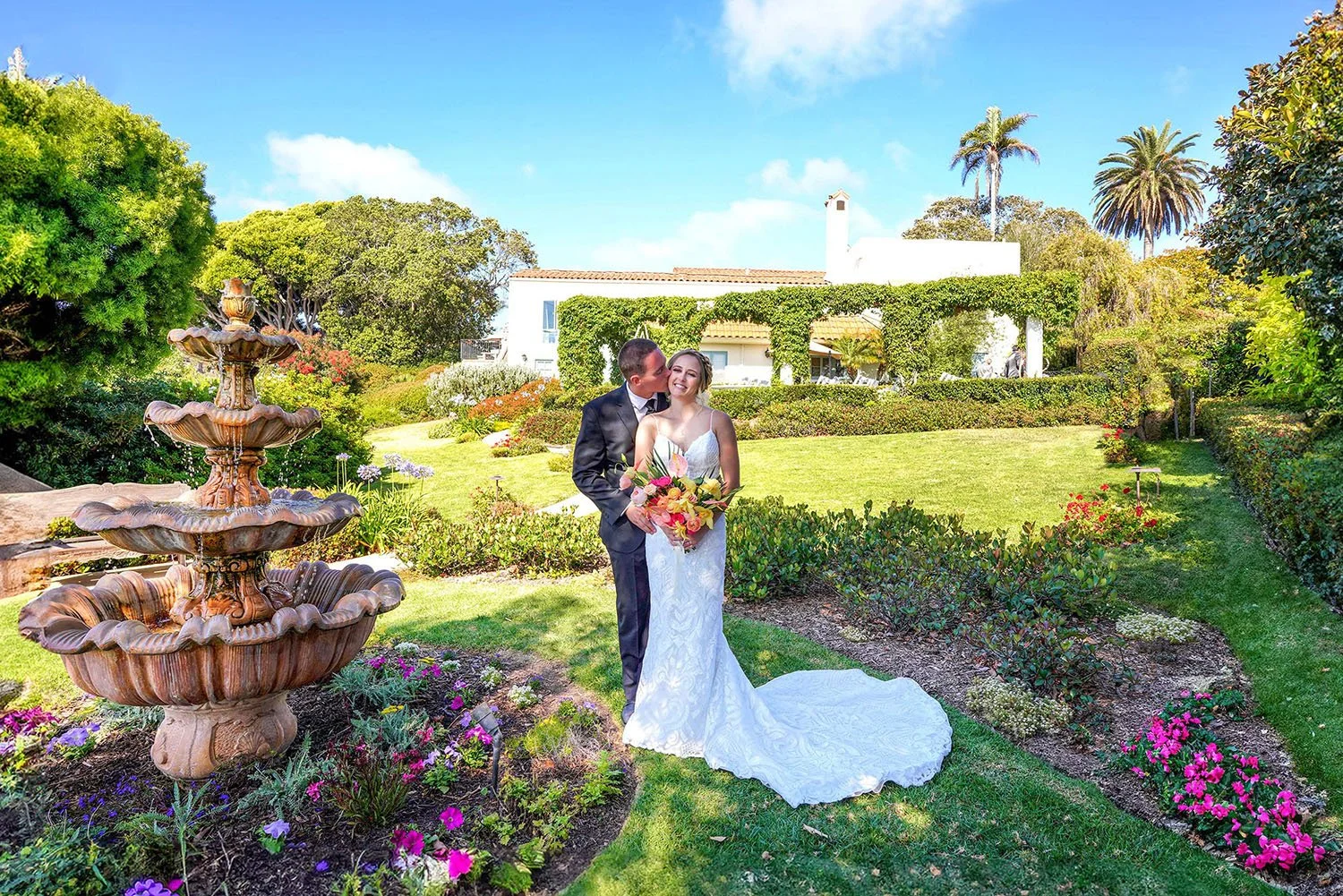A smiling bride and groom stand in a lush, sun-drenched garden at the Thursday Club in San Diego's Point Loma.
The groom, in a dark suit, kisses the bride's cheek as she holds a vibrant bouquet of tropical flowers in shades of pink, orange, and yello