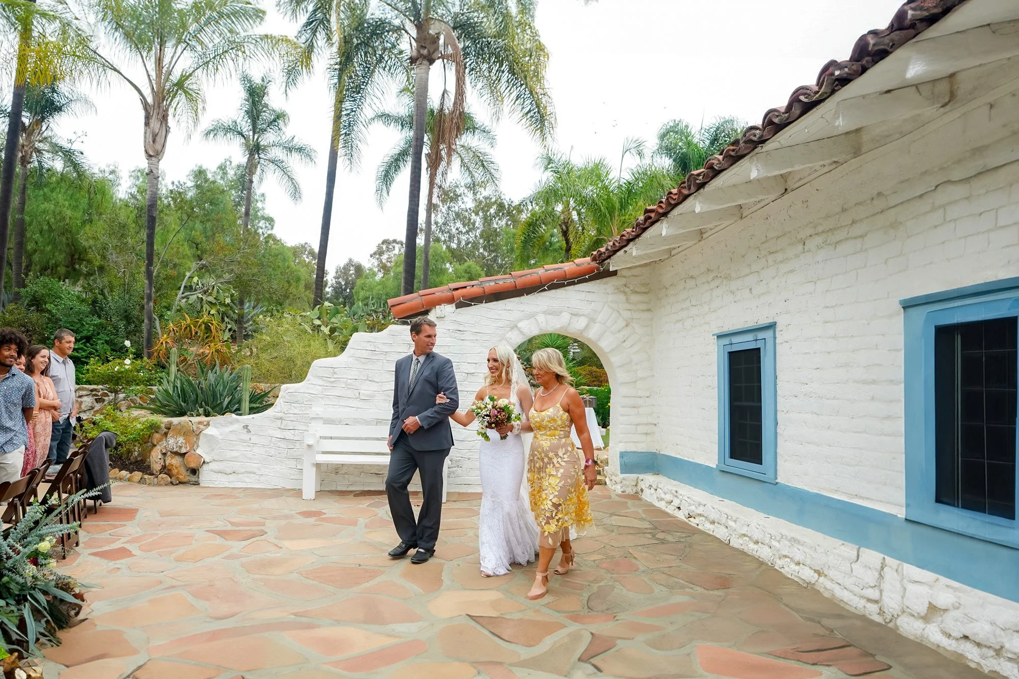 A photo of a bride in a white lace dress and veil walking down an outdoor flagstone aisle, escorted by a man in a dark grey suit and a woman in a yellow floral dress. They are passing a white hacienda-style building with blue-trimmed windows and a st