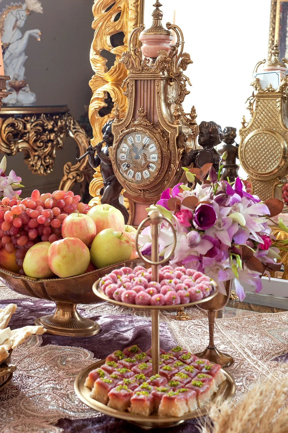 An elaborate, formal table setting, likely a Persian wedding Sofreh Aghd, featuring an antique gold clock, a two-tiered stand with pink and green sweets and pastries, a bowl of red grapes and green apples, white and purple floral arrangements, and or