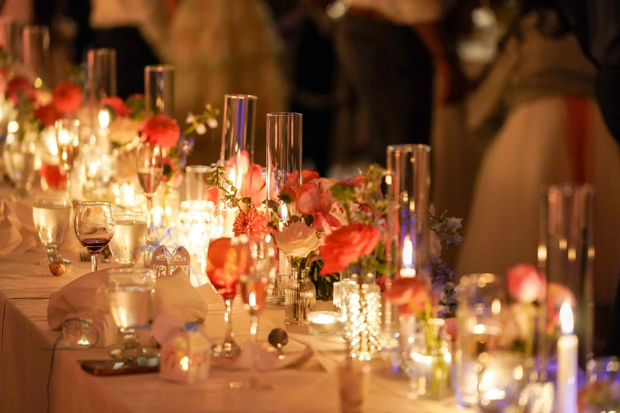 A beautifully decorated long dinner table set for a formal event or wedding reception, illuminated by many flickering candles in clear glass holders and adorned with arrangements of pink, white, and red flowers and various glassware.