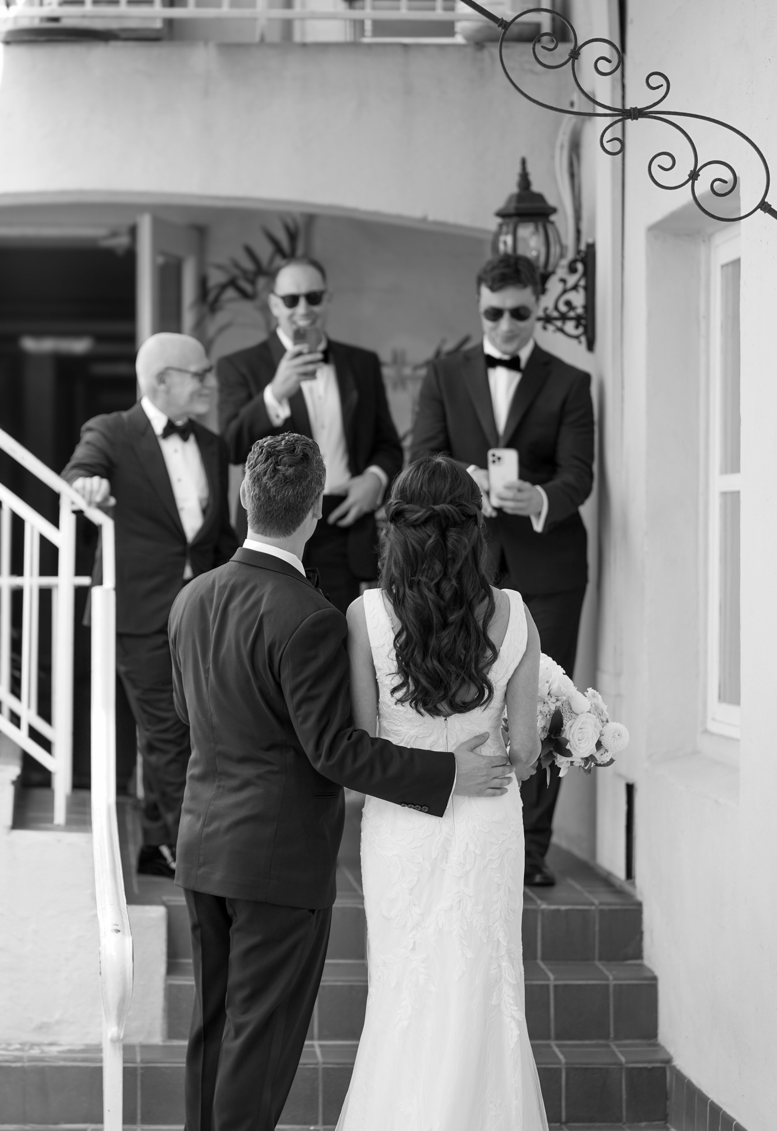 Candid black and white photo from behind a bride in a lace gown and a groom in a tuxedo as they walk toward guests taking photos on their phones.