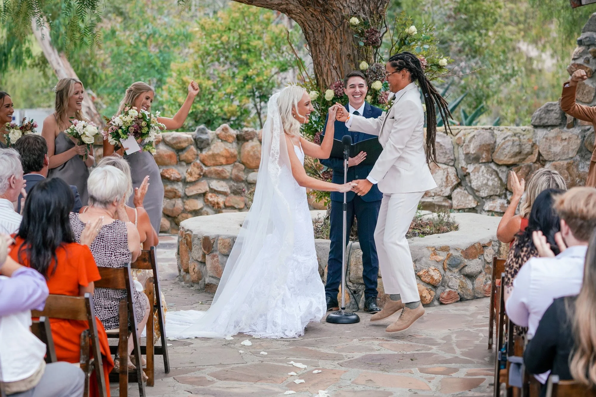 A photo of two brides joyfully dancing together on a flagstone patio after their wedding ceremony, surrounded by clapping guests and bridesmaids.


