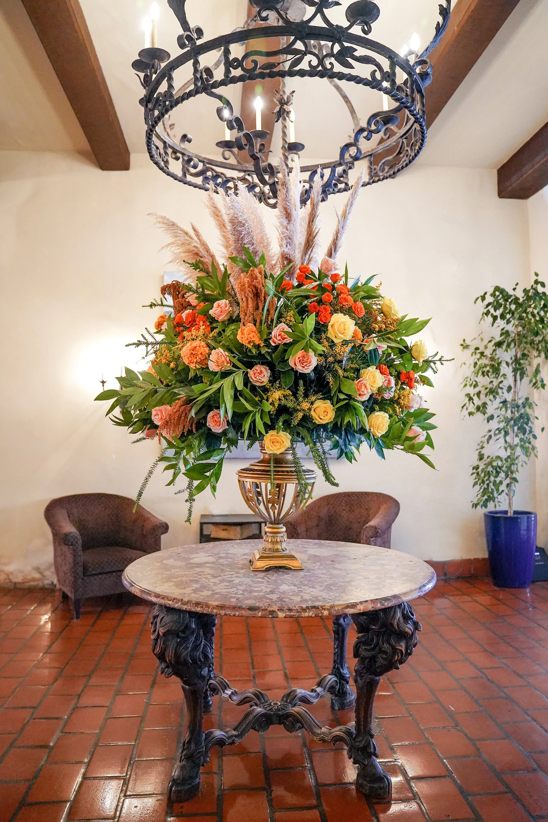 A round, marble-topped table with a heavily carved, dark wooden base stands in the foreground. Centered on the table is a decorative, gold-toned urn overflowing with a large, vibrant floral arrangement featuring orange, yellow, and peach roses, as we
