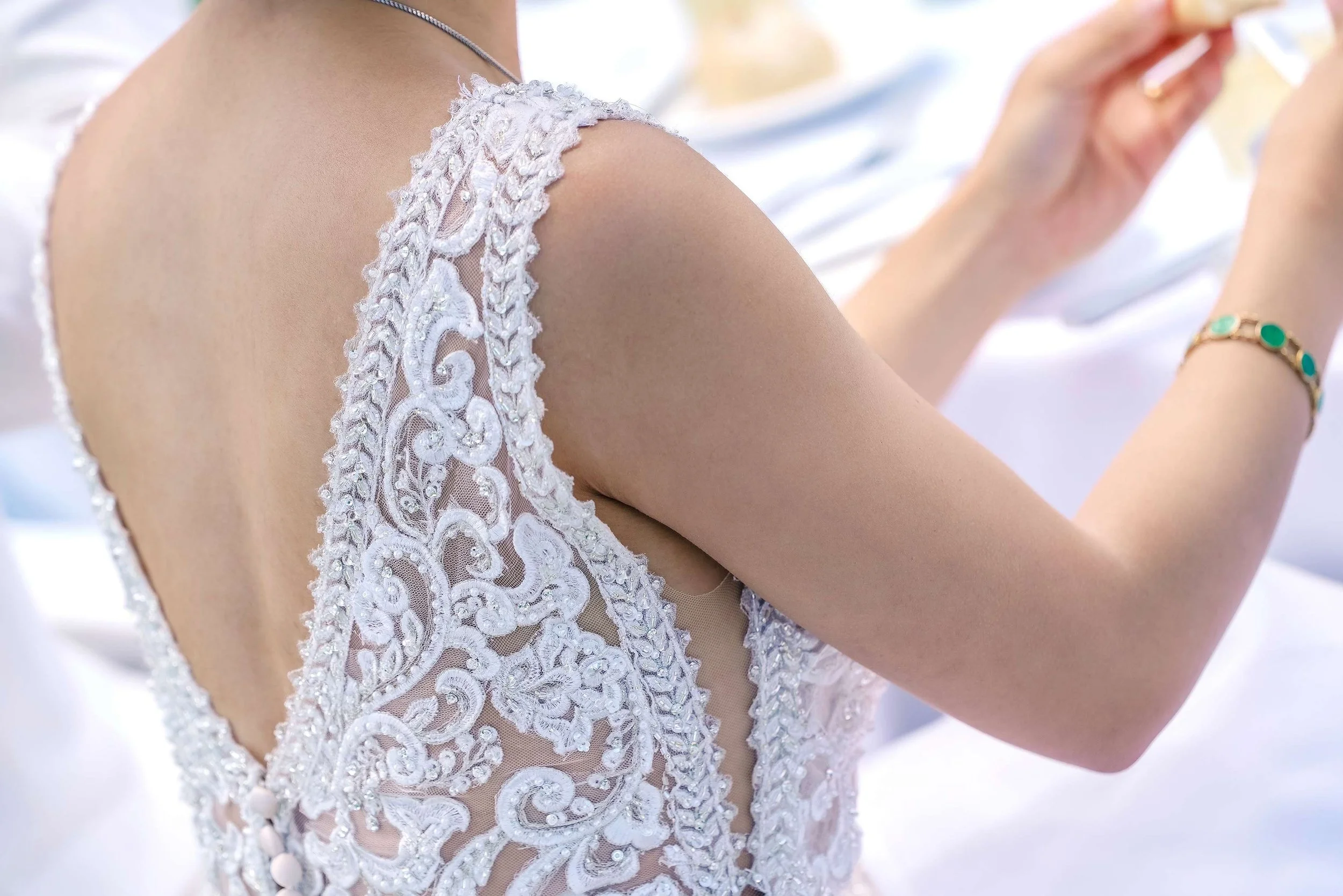 A close-up shot of a bride's back, showcasing the intricate white lace appliqué and sheer paneling of her wedding dress with a deep V-neckline as she is seated at a table.