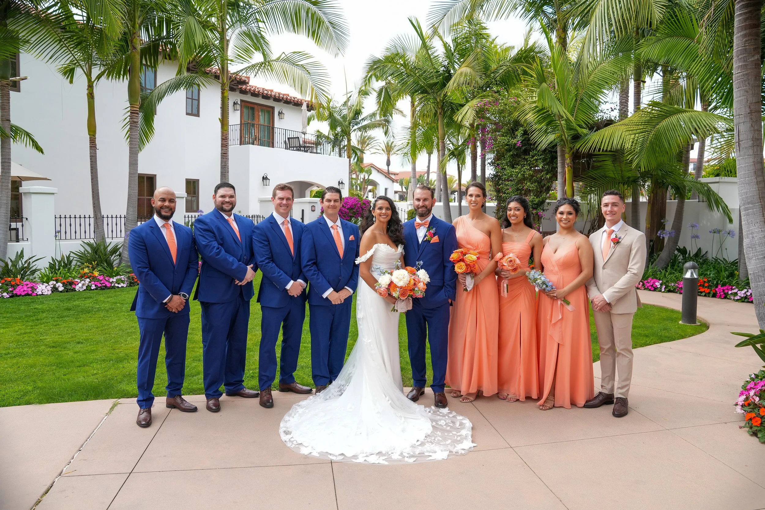 A wedding party portrait by Reimo Photography at Omni La Costa Golf Club, featuring a bride and groom standing with their wedding party on a paved path lined with palm trees. The groomsmen wear bright blue suits with coral ties, the bridesmaids wear 