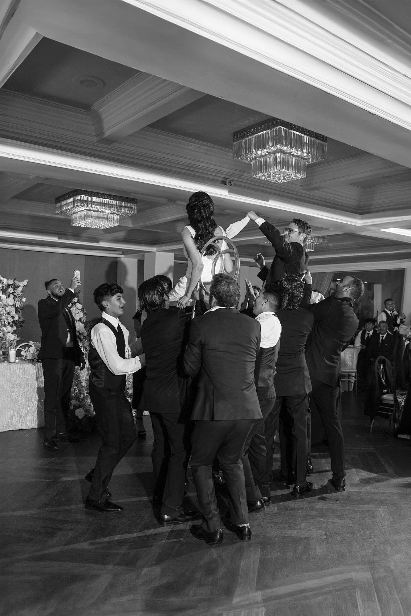 A black and white photograph of an indoor wedding reception where a person in a white outfit is lifted in the air on a chair by a group of men in suits. The people are on a dark wooden dance floor, and the room has decorative chandeliers and other gu