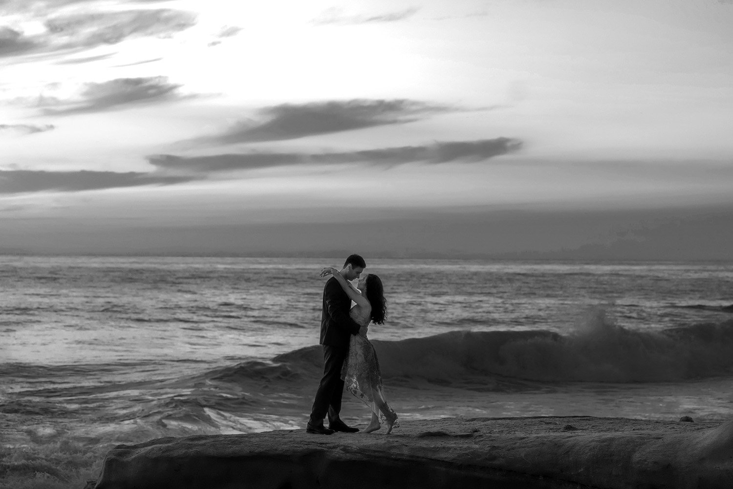 An outdoor photograph of a couple embracing on a large rock formation by the ocean during a beautiful sunset or sunrise. The man is wearing a dark suit, and the woman is in a flowing, patterned dress. The sky is filled with warm orange, pink, and pur